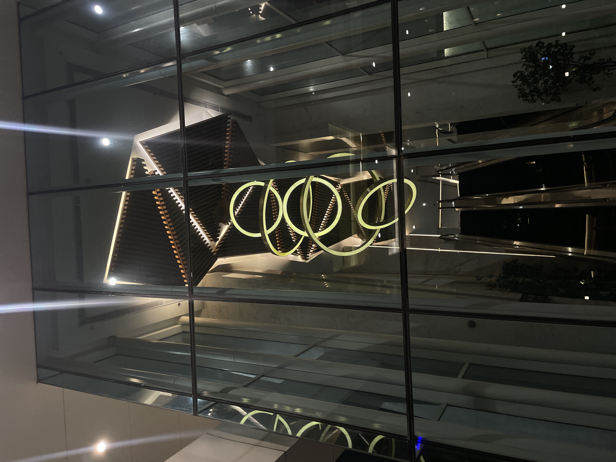 The image captures the interior of a modern building, likely a hotel lobby or a commercial space, viewed from the exterior through a large glass facade. The scene is dimly lit, with the primary light sources being elegant, circular pendant lights that hang in a cascading arrangement in the center of the atrium. These lights are a soft yellow hue and create a striking visual focal point against the darker backdrop.
The architecture features clean lines and geometric shapes. Above the pendant lights, a multi-tiered ceiling structure with wooden slats and integrated lighting is visible, adding a sense of depth and sophistication. The walls appear to be clad in a light-colored material, possibly marble or a similar stone, which reflects the ambient light.
In the lower portion of the image, a set of escalators is visible, suggesting the building's functionality as a multi-level space. The escalators are dark in color, with metallic trim, and appear to be in operation or ready for use.
Reflections on the glass facade create a layered effect, showing the interior elements superimposed on hints of the exterior environment, though the exterior is largely indistinct due to the low light conditions. The overall impression is one of contemporary luxury and a well-designed, atmospheric interior space. The building's facade is divided into a grid of large glass panes framed by dark metal mullions.
