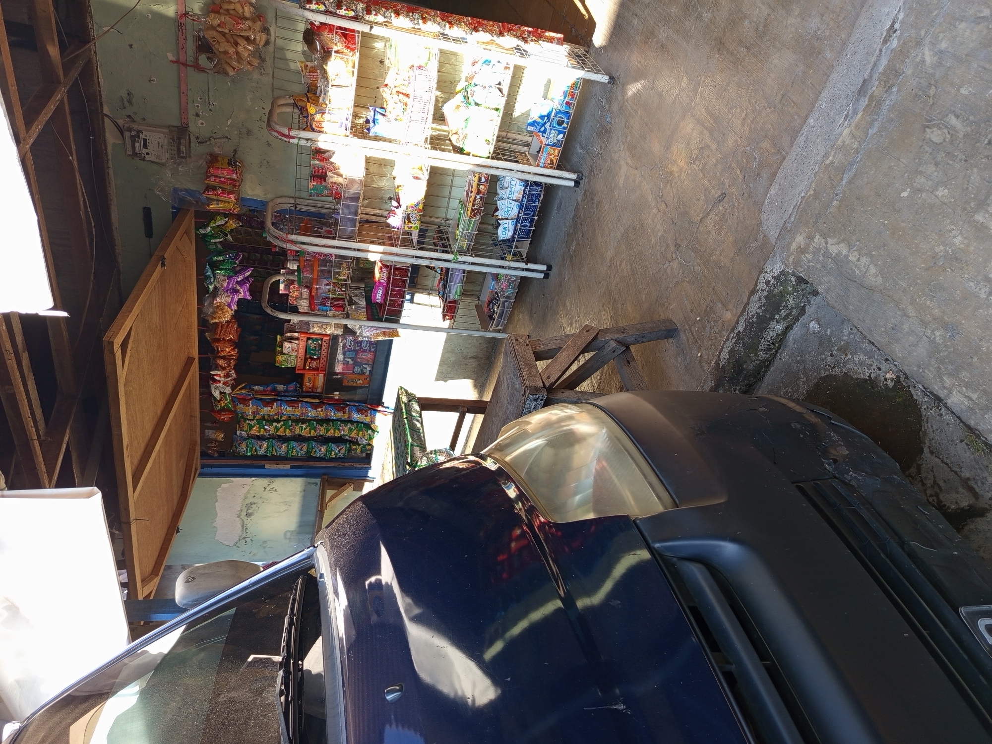 A dark blue van is parked in front of a small, open-air convenience store. The store has shelves stocked with various packaged goods, including snacks and drinks. A wooden stool and a metal rack are also present in front of the store. The ground is concrete, and sunlight is casting shadows. The location is identified as Tebel, Indonesia.