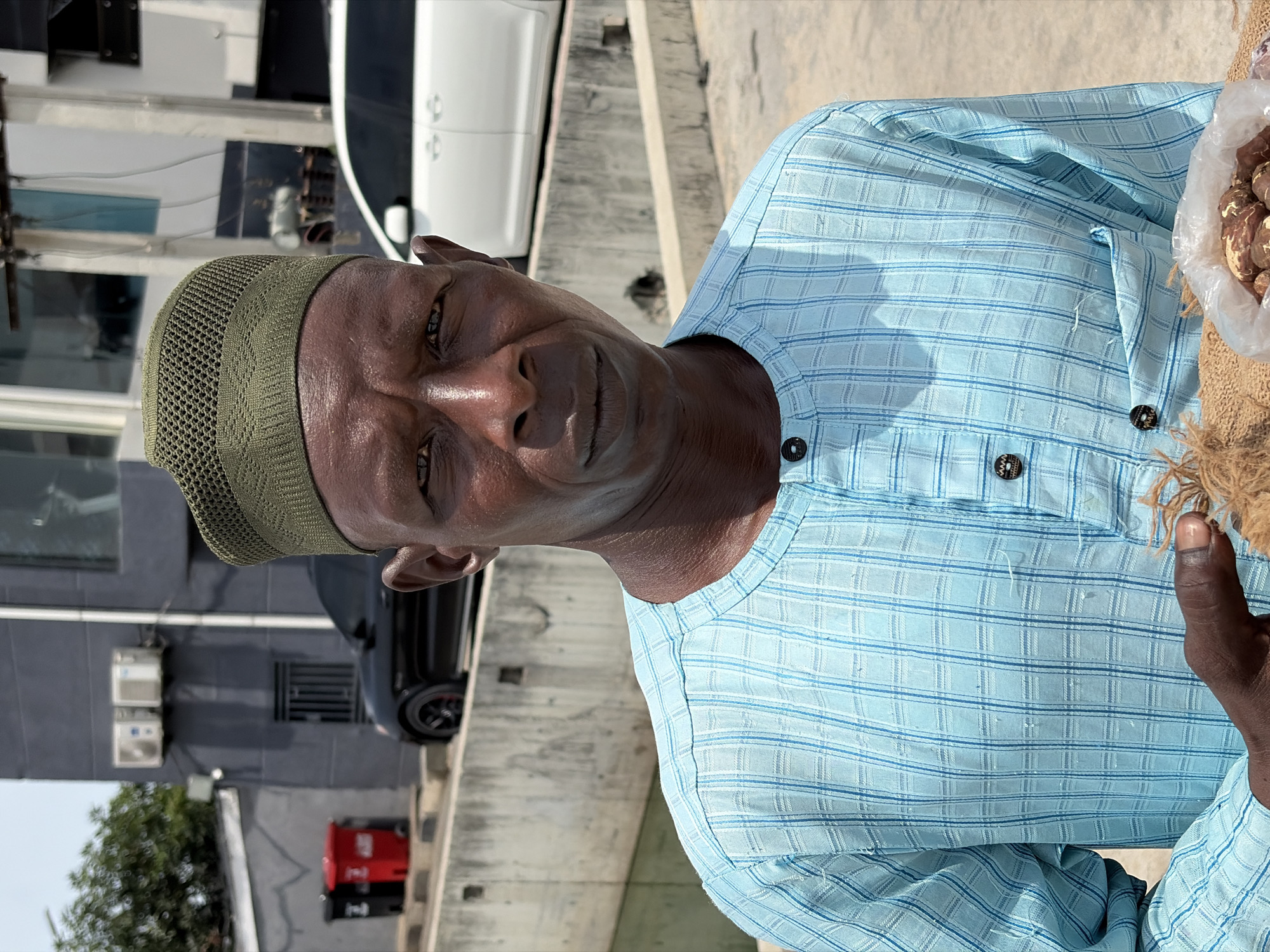 A man with dark skin and a khaki crocheted skullcap is wearing a light blue checkered shirt with three buttons. He stands in front of a grey building in Igboefon, Nigeria with air conditioning units on the wall. He is holding what appears to be a bag of nuts. There is a black car and white car in the background. A red generator is visible by the building.
