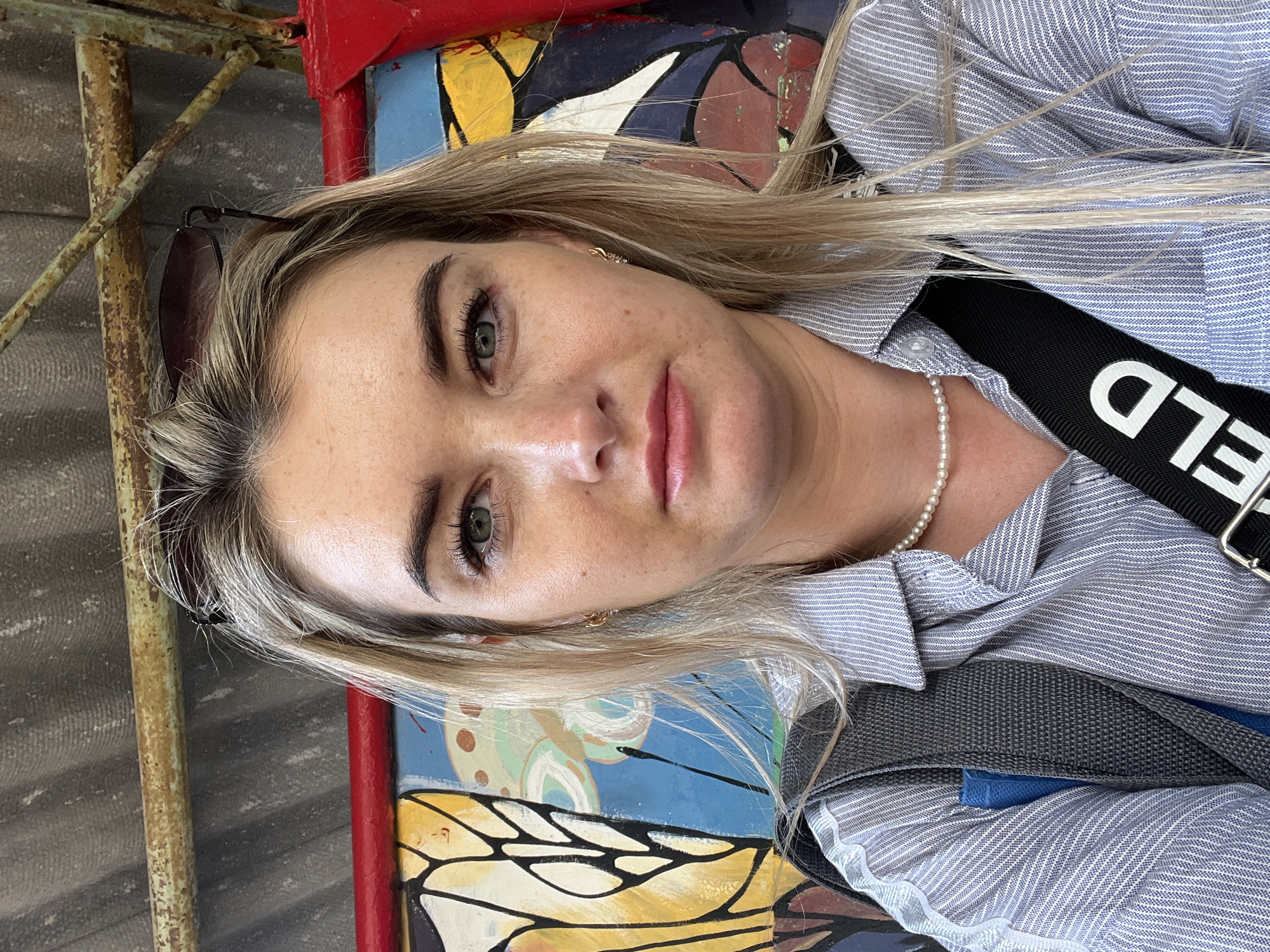 A blonde woman is captured in a close-up selfie. She has fair skin, green eyes, and lightly pink lips. She is wearing a light blue striped shirt, a pearl necklace, and a black strap bearing white letters. Sunglasses are perched on her head. The background is a painted mural featuring colorful geometric shapes and a butterfly design. To the left, there is a rusty metal structure. The photograph appears to be taken in Chaikovsky, Russia, based on information associated with the source image.