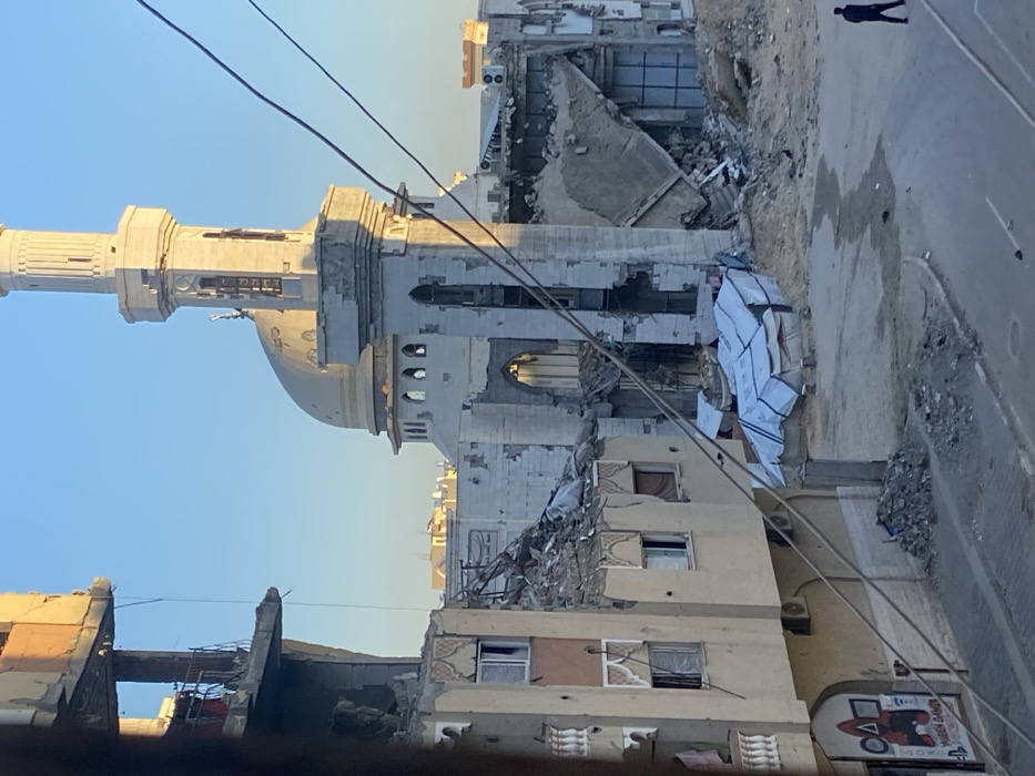 The image depicts a heavily damaged building, potentially a mosque, with a partially collapsed minaret. Debris and rubble are scattered around, indicating significant destruction. A person walks along the street adjacent to the building. Power lines stretch across the foreground, and the sky is clear. A partially destroyed adjacent building, perhaps residential, is visible. The overall scene evokes a sense of devastation and loss.