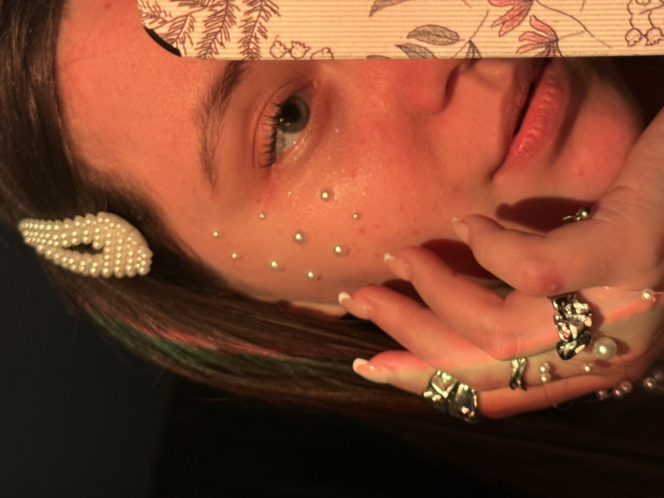 A close-up image depicts the right side of a female subject's face and a portion of her left hand, illuminated by warm, reddish-orange light. The subject possesses dark brown hair with visible streaks of green and red, secured above the right temple with a white, pearl-embellished bobby pin. Approximately twelve to fifteen small, spherical, white beads are adhered to the right cheekbone and below the right eye. The visible right eye is blue or grey-blue. The left hand, positioned near the right cheek, displays a French manicure on the fingernails and features three silver-toned rings: one with an organic, textured design on the index finger, another with a crossed design on the ring finger, and a third with a pearl or light-colored stone on the pinky finger. A delicate strand of small white pearls is visible around the lower part of the left hand. The right side of the subject's face, including the nose and mouth area, is partially obscured by a vertically held, light-colored, rectangular object adorned with dark botanical patterns, including leaves and stylized flowers. The background is indistinct and dark. The scene's precise location within the city of Krasnodar, Russia, cannot be determined from visual evidence in the image.