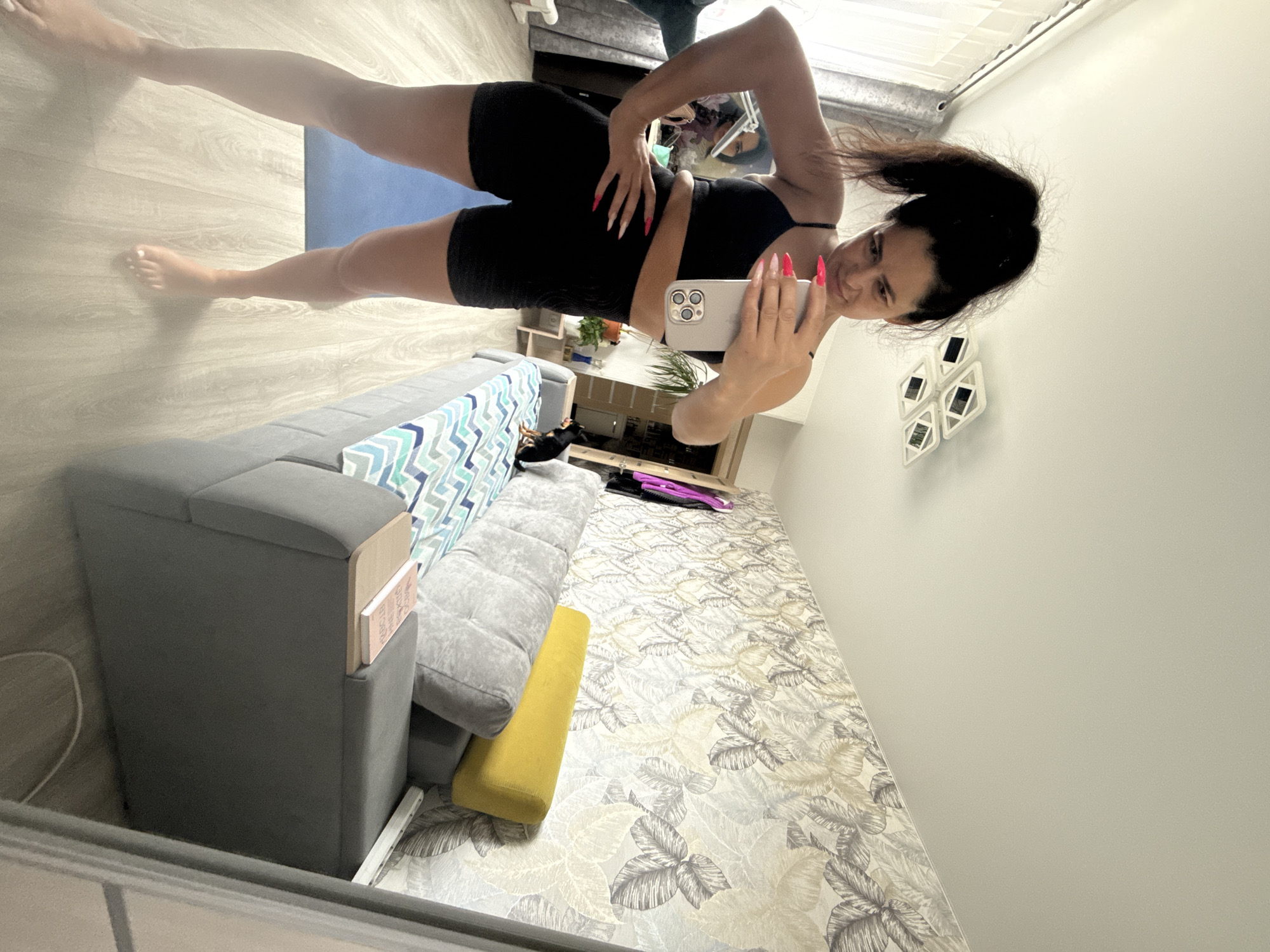 A woman is taking a selfie in an interior space. She is wearing a black sports bra and black shorts. She is standing on a wooden floor, partially covered by a blue yoga mat. A gray sofa with blue and white cushions is visible in the left foreground. A printed rug lies on the floor. Various objects such as books, plants, and other furniture items are also in the scene. The location is Kauchuk Stantsiya, Russia.