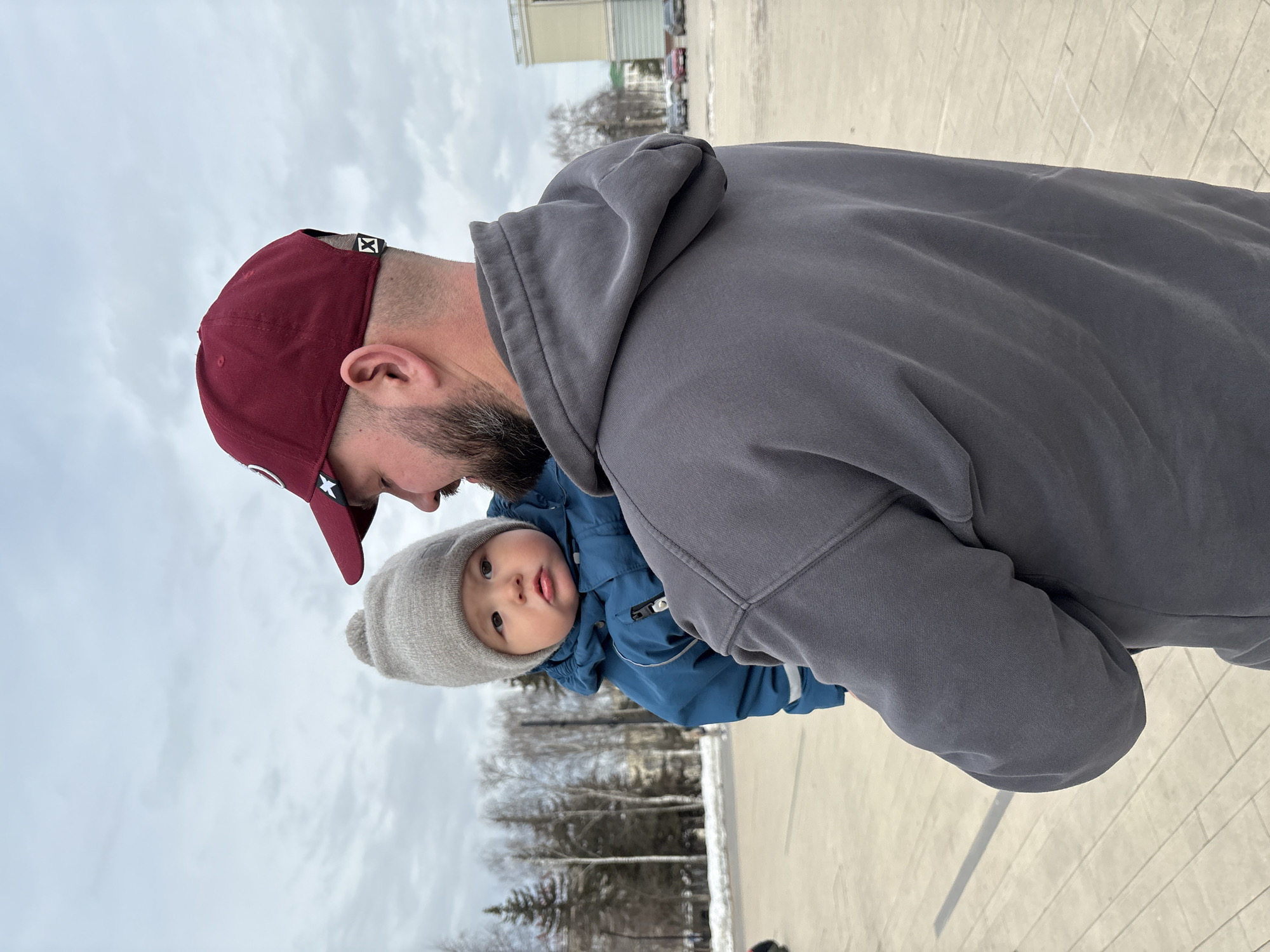 A man wearing a maroon baseball cap and a gray hoodie holds a baby in a blue coat and gray beanie. The baby is sticking their tongue out. The background shows trees, a building, and a cloudy sky. The location is a paved area in Izhevsk, Russia.