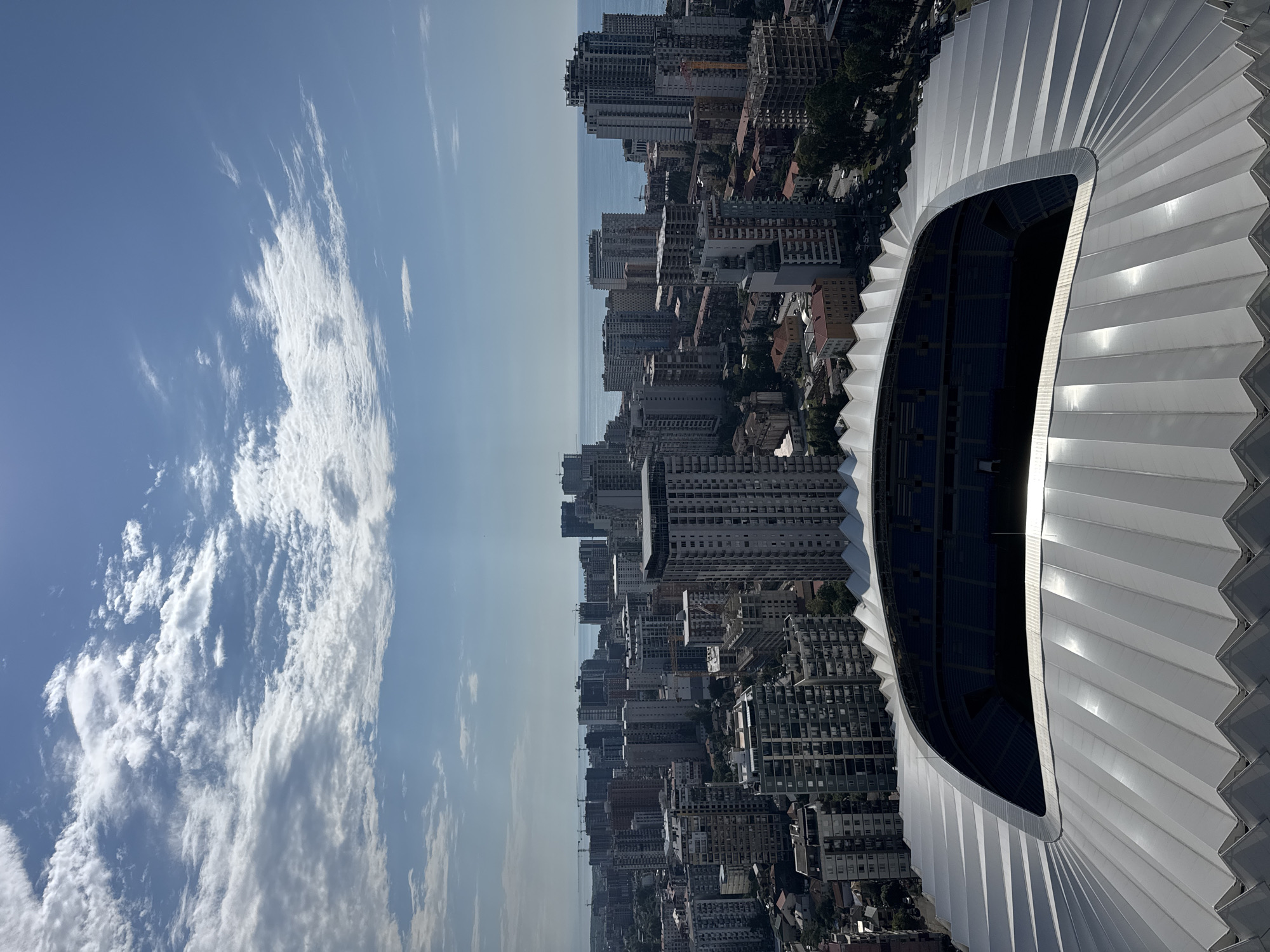 An aerial, high-angle shot looks down upon the city of Batumi, Georgia. In the foreground is the stadium roof of the Batumi Arena, a large, modern structure with a white, ribbed canopy that curves around the seating area, which is visible as blue rows of seats. Beyond the stadium, a dense urban landscape of tall, modern apartment buildings stretches towards the horizon. The buildings vary in height and architectural style, with many featuring balconies and glass facades. In the distance, the Black Sea is visible, its blue expanse meeting the sky. The sky is a bright blue, with scattered white clouds, some of which are wispy and thin, while others are more dense. Sunlight shines brightly, casting shadows and illuminating the scene. The overall impression is of a sunny day in a coastal city with significant development.