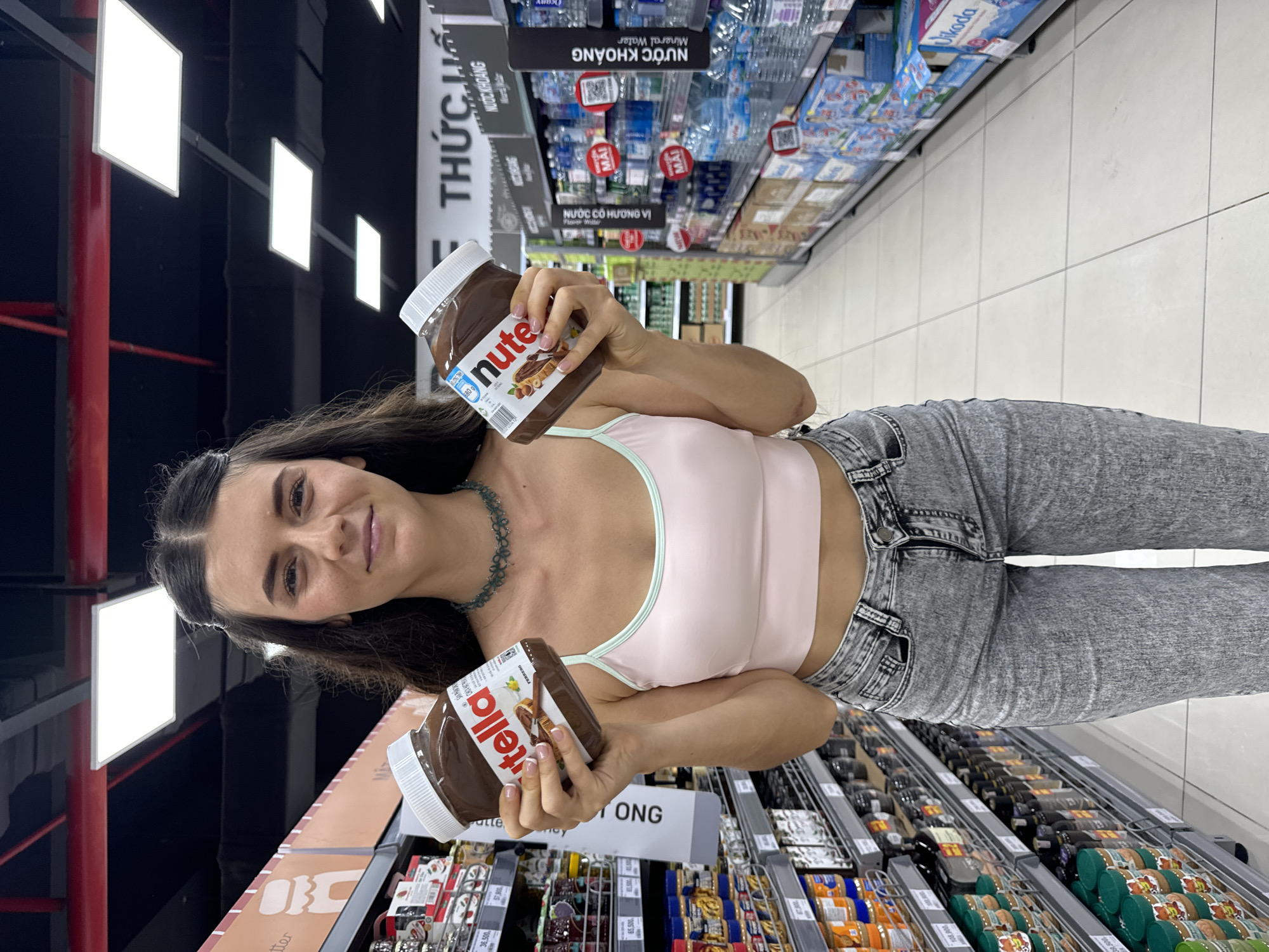 The image captures a woman standing in the Phu Thuy Ward, Vietnam supermarket aisle. She is holding two jars of Nutella. She wears a light pink tank top with thin straps, and light-wash gray jeans. Her dark hair is pulled back from her face. She is smiling. Supermarket shelves filled with various products are visible behind her. Aisle signage, including "THỨC UỐ" and "NƯỚC KHOÁNG," is also visible. There are overhead lights on the dark ceiling.