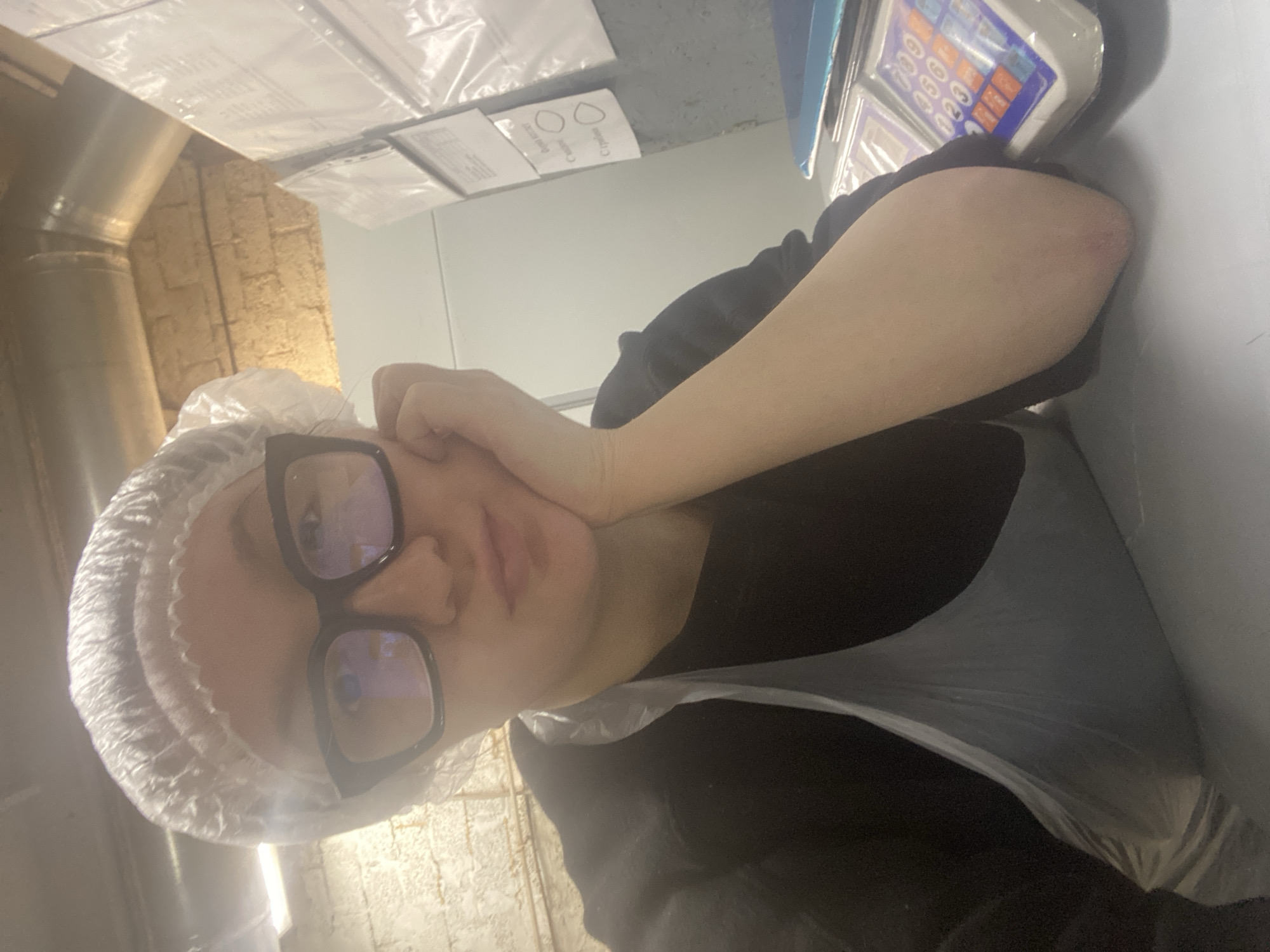 A person with a plastic hairnet and black-rimmed glasses rests their face on their hand. They are wearing a black garment over which a clear plastic apron is placed. The background features an industrial-style metal pipe, a brick-like wall, and a light-colored wall with papers attached. A weighing scale is visible in the upper right corner. The location is likely a food preparation area, possibly in Chaikovsky, Russia, based on the request.