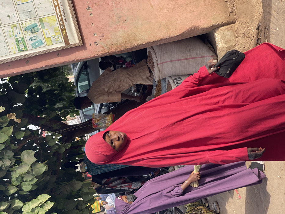 The image depicts a bustling outdoor market scene in Samaru, Nigeria, illuminated by direct sunlight. In the foreground, a woman wearing a vibrant red full-body abaya/hijab is positioned centrally, her face visible and looking towards the right of the frame. Her right hand holds a small black pouch or cap featuring a white Nike logo. Faint dark markings, consistent with henna or tattoos, are visible on her exposed right forearm. To her left, slightly behind her, another woman in a purple full-body abaya/hijab and a black face mask holds a bundle of light-colored sticks in her right hand.
On the far left, a pile of green and yellow bananas rests on the ground. Above, a large tree with dense green foliage provides shade, and various fabrics or garments are displayed hanging from a stand beneath its branches, alongside small packaged items suspended by strings. In the mid-ground, a male figure dressed in a light beige caftan is partially obscured, appearing to attend to several large white sacks on the ground, which are filled with light-colored grains or nuts. These sacks are situated alongside a textured, faded reddish-pink wall that forms the right boundary of the image. Affixed to this wall is a white rectangular sign displaying various cosmetic products and text including "TEL: 08107222094, UR BEAUTY". The background reveals parked vehicles on a street and additional blurred individuals, indicating a crowded urban environment.