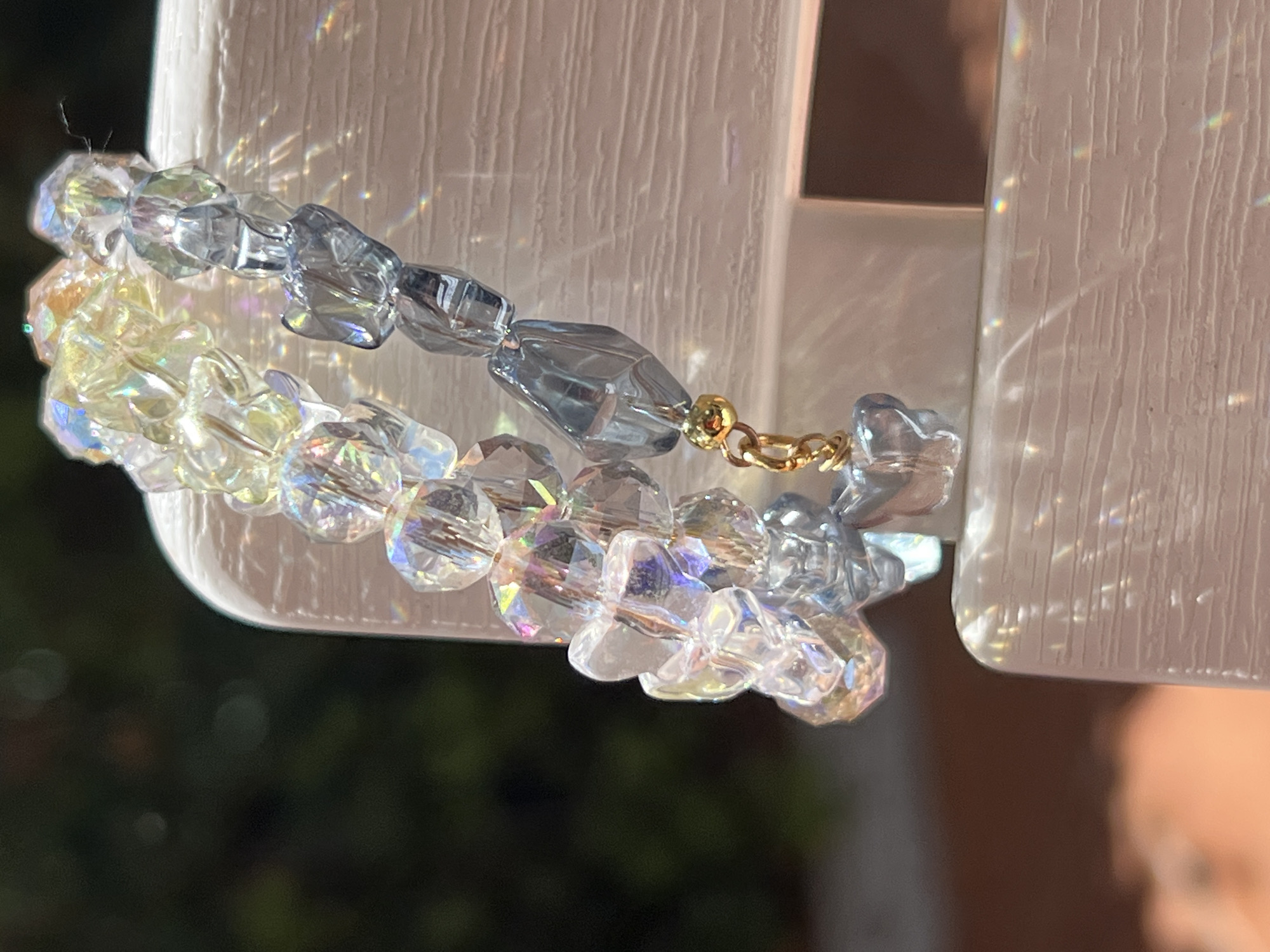 A close-up, eye-level shot focuses on two strands of iridescent and translucent blue glass beads, strung together with a gold clasp. The beads vary in shape and size, with some appearing faceted and others smooth and rounded. They are draped over a textured, white, possibly wooden surface that reflects light, creating rainbow-like refractions and glare across the image. The background is blurred, showing dark green foliage and a hint of reddish-brown. The scene is lit with strong, directional light, suggesting it is outdoors or near a window. The image does not contain information about the city of Bijela, Montenegro.