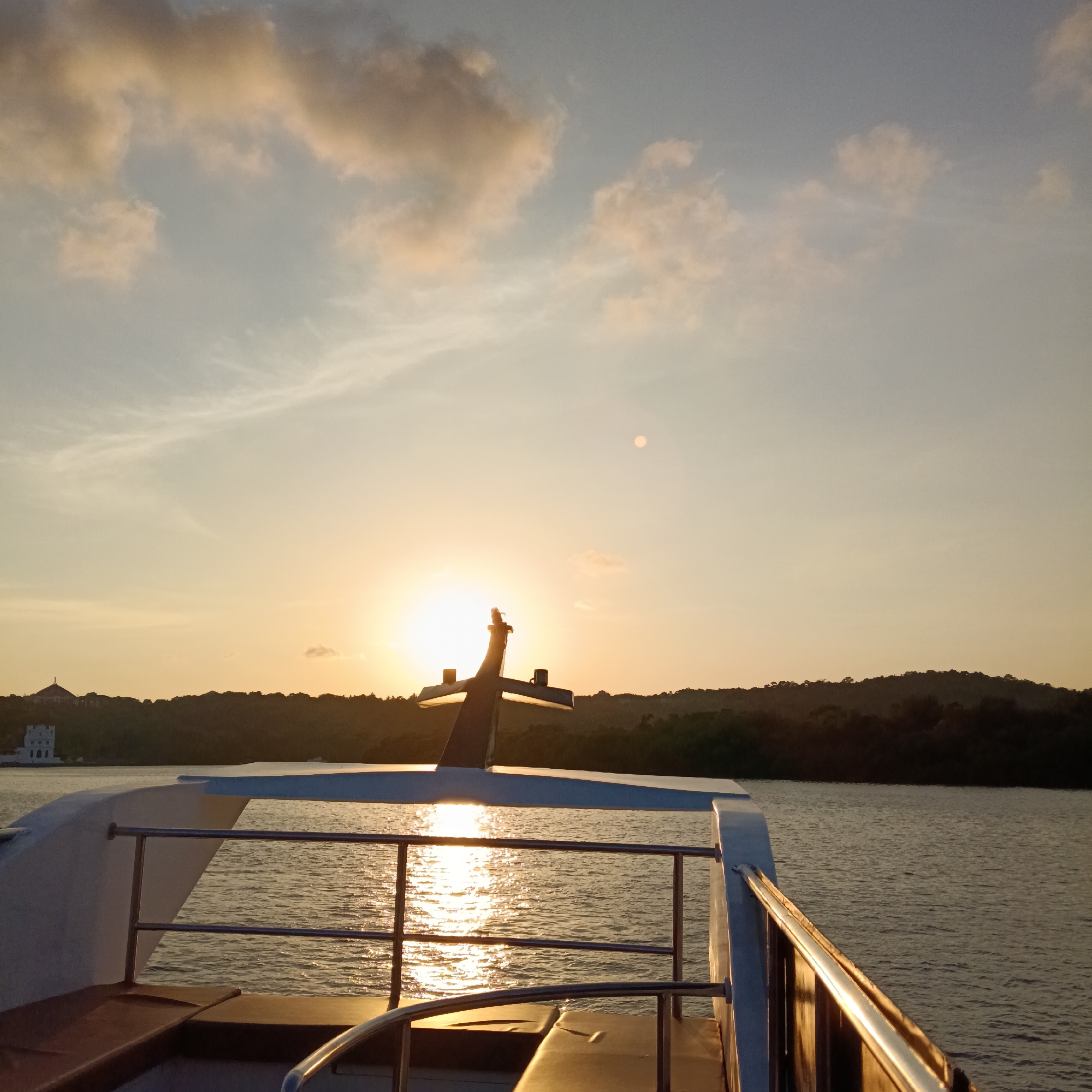 The image shows a view from a boat on a body of water, presumably a river or estuary, with the sun setting in the background. The location is in or near Britona, India.
In the midground, a large cross-shaped structure is silhouetted against the setting sun. The structure is part of the boat itself. Sunlight is reflected on the water surface.
The boat's railing and deck are visible in the foreground. In the background, a tree-covered shoreline can be seen with a white building that is possibly a church. The sky is light with clouds.