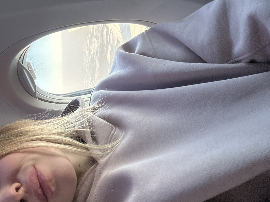 A blonde female is depicted in a close-up, seated inside an airplane cabin during daytime flight. Her lower face, including her mouth and chin, and light blonde hair are visible on the left side of the frame. She wears a plain, light lavender-colored hooded sweatshirt. To her right, an oval airplane window reveals an aerial view of an extensive layer of white clouds below, beneath a clear light blue sky. A portion of the airplane's wing, light in color, is visible at the lower right edge of the window. The window frame is a light gray with a small operating latch at the top. The scene depicts an in-flight journey, contextually associated with Teigebyen, Norway.