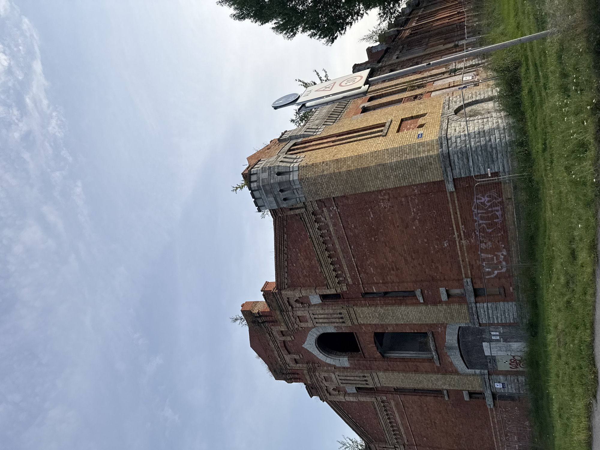 The image shows a section of the Kalamaja Barracks in Tallinn, Estonia. The barracks are constructed of red brick with yellow brick accents and stone foundations. The building appears to be abandoned or undergoing renovation, with boarded-up windows and doors, graffiti on the lower sections, and temporary fencing. Several trees are visible around the building, and the sky is partly cloudy. A street signpost with traffic regulations is also present. The scene is captured from a low angle, looking up towards the building.