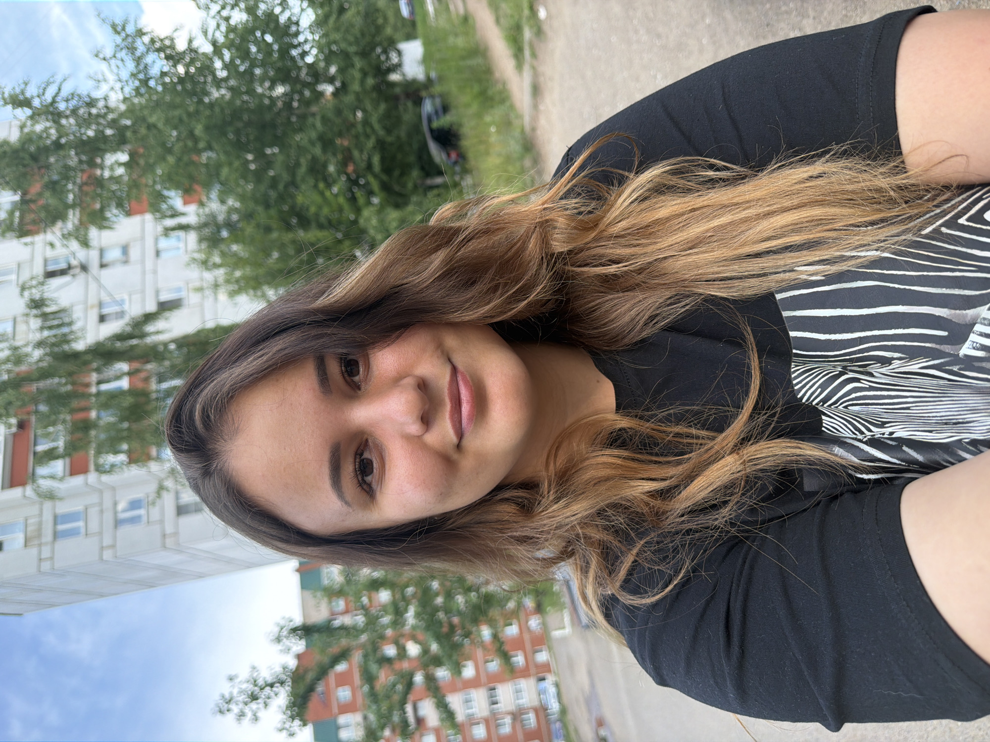 In Chaikovsky, Russia, a young woman with shoulder-length brown hair, lighter at the ends, smiles at the camera. She is wearing a black t-shirt with a white zebra-striped pattern on the front. Behind her, a tall, light-colored apartment building with red accents is visible, along with green trees and a cloudy sky. The ground appears to be a paved area, possibly a courtyard or parking lot.