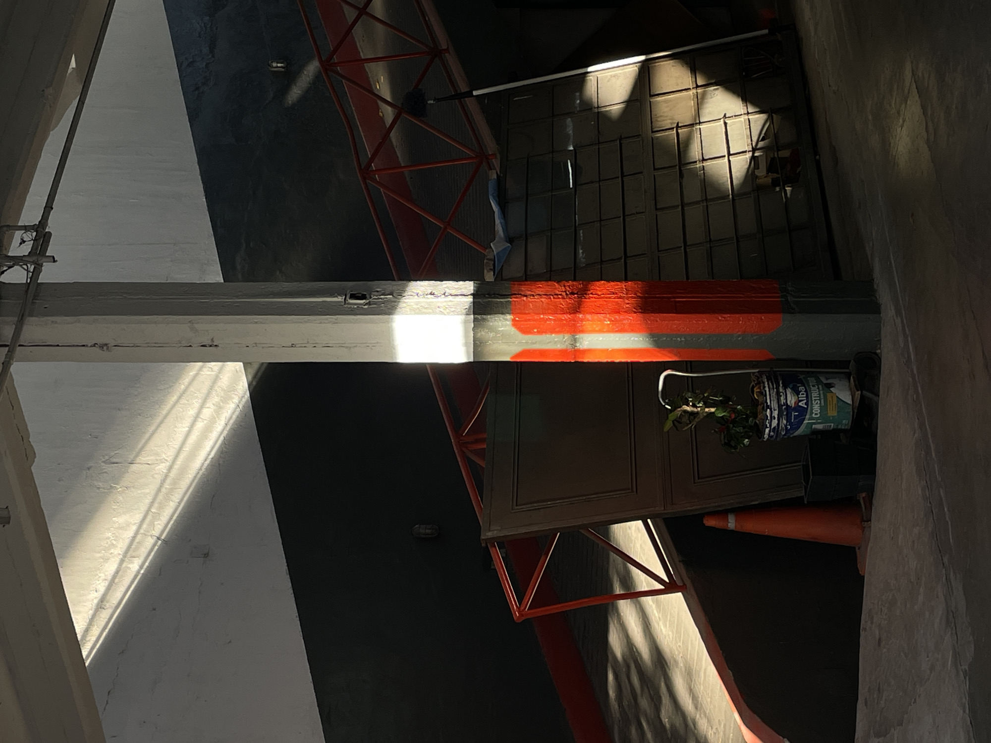 The image depicts an architectural or industrial interior space, presented in a portrait orientation that suggests a view looking up or down into a shaft or complex structure. A prominent, light grey concrete pillar extends vertically through the frame, marked by a bright orange band of paint on its lower right section. Intense sunlight illuminates portions of this pillar and an adjacent white wall, casting sharp, angular shadows across the scene and creating stark contrasts with darker areas.
To the right of the central pillar, a red, angular metal structure, possibly scaffolding or a support frame, is visible. Below this, a dark, grid-like metal surface is partially seen, also patterned with strong shadows. In the lower right, an orange traffic cone stands next to a dark brown door or panel. A white paint bucket containing a small plant rests on the ground near the door; legible text on the bucket includes "Alba Constructora" and "Pintura." The background comprises dark grey and black walls or surfaces, some displaying texture. No people or discernible activity are present. The distinct, high-contrast lighting indicates daytime with clear weather, likely morning or late afternoon. The visible text "Alba Constructora" is a notable detail, aligning with the specified location context of Buenos Aires, Argentina.