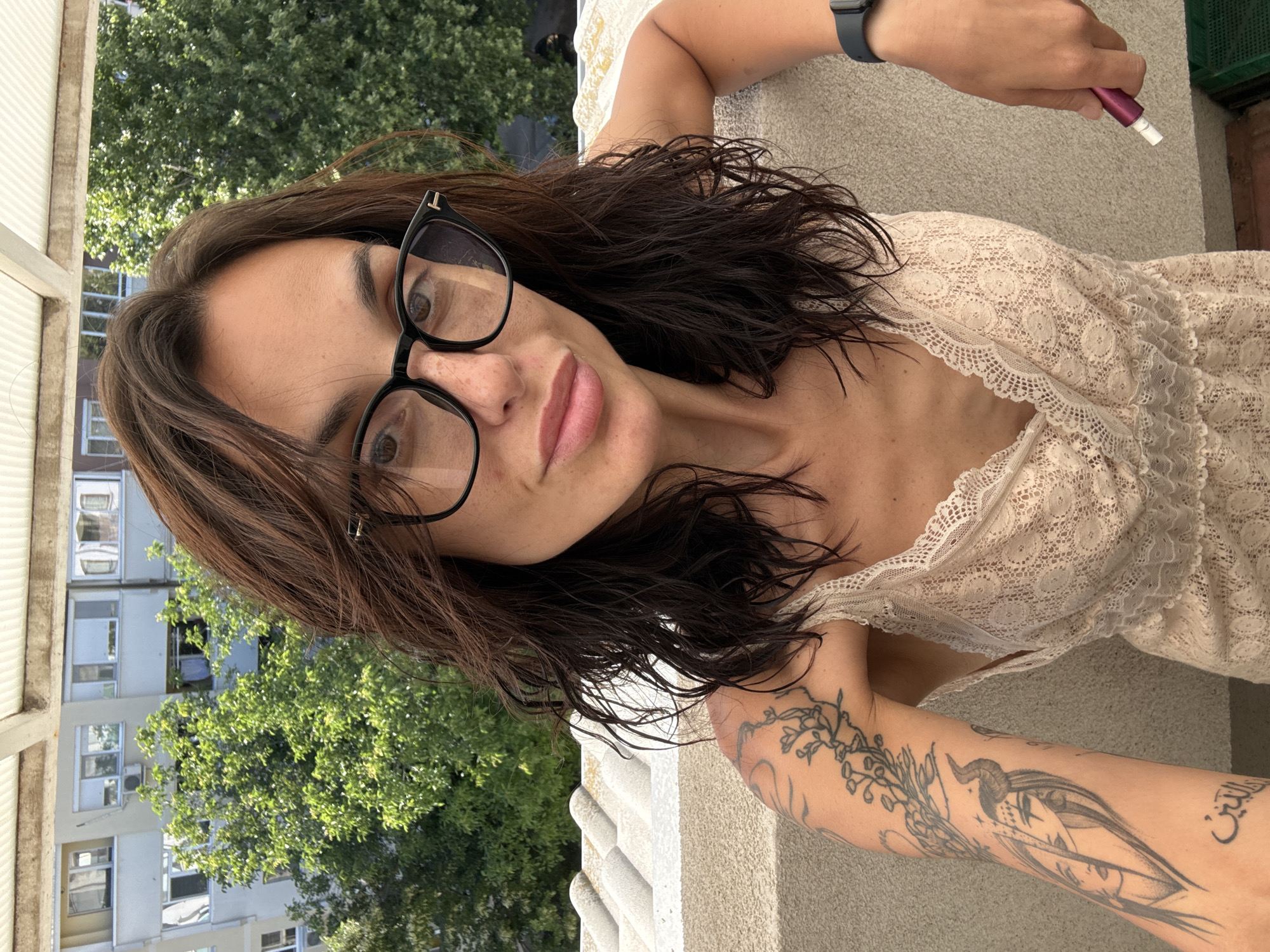 A woman with long, dark, wavy hair and glasses is taking a selfie on a balcony in Pleven, Bulgaria. She is wearing a cream-colored, lace-trimmed dress and has tattoos on her left arm. The background shows trees and apartment buildings, indicating an urban setting. She is holding a pink object in her right hand.