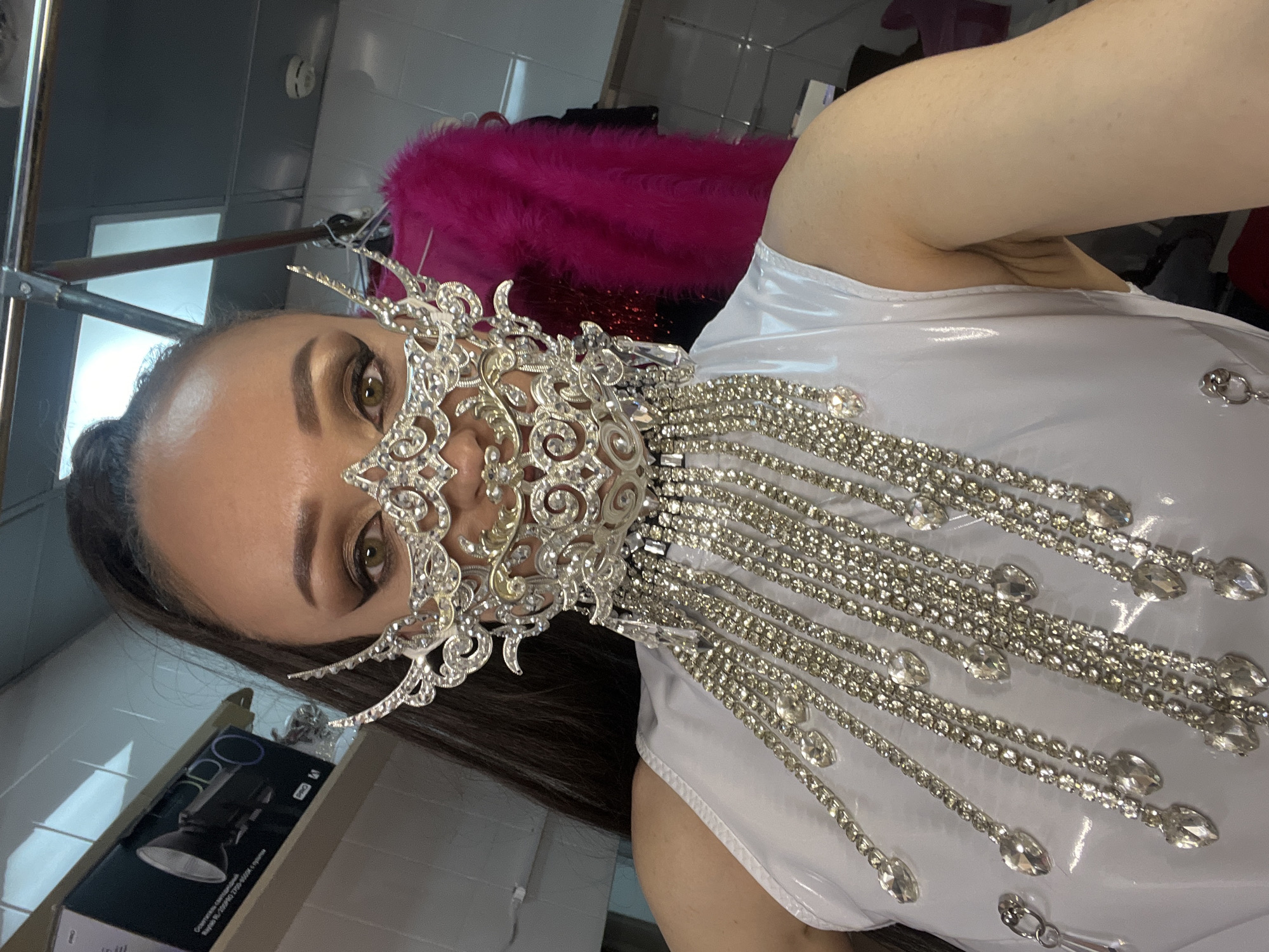 A woman is shown in a close-up selfie, wearing an elaborate silver mask that covers the lower half of her face. The mask features intricate designs and long, dangling chains with gem-like adornments. She is wearing a shiny white sleeveless top, also embellished with similar gem-like details.
The location appears to be indoors, possibly a dressing room or backstage area, given the presence of clothing racks and other items visible in the background, including a pink feather boa or similar garment. A box is also visible in the bottom left corner of the image.
While the image does not contain direct evidence to confirm the location in Kazan, Russia, it can be determined that the location of this photograph cannot be determined based only on these objects.