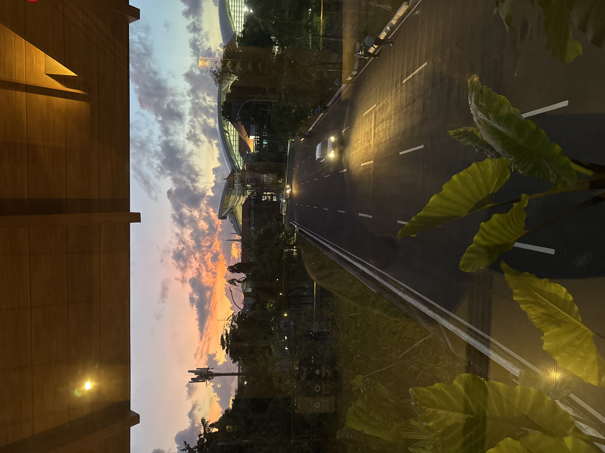 Here is a detailed description of the image:
The image presents a nighttime scene from an elevated perspective, likely from a window or balcony. The view is dominated by a multi-lane road illuminated by car headlights and streetlights. The road runs through a city with visible buildings, trees, and infrastructure.
The left side of the frame features a structure, perhaps a building facade, with a warm, artificial light reflecting off its surface. In the background, a colorful sunset or twilight sky is visible, with hues of orange, pink, and blue blending together. Clouds add texture to the sky.
On the right side, there are large green leaves, suggesting the presence of a plant either inside the building or very close to the window. The leaves overlap parts of the road scene, adding a sense of depth to the image.
The location in the city is Unknown, Unknown, but the presence of a stadium-like structure with distinctive architectural features can be seen in the background, indicating a major urban area.
