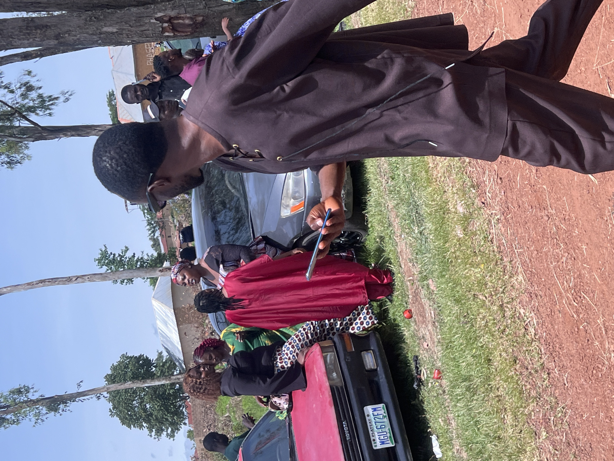 The image depicts an outdoor scene in Wumat, Nigeria, where a group of people are gathered near several vehicles. In the foreground, a man wearing a dark brown, loose-fitting garment walks away from the camera, holding a smartphone. Behind him, a dark sedan is visible. To the left, a red Toyota car is parked, with a license plate reading "PLATEAU MGU-672FM". A woman in a black top and patterned pants is leaning on the hood of the red car, interacting with other individuals. A woman in a bright red garment stands near the red car, facing away from the camera. Further back, other people are seen standing around, with buildings and trees forming the backdrop under a clear sky. The ground is a mix of grass and dirt.
