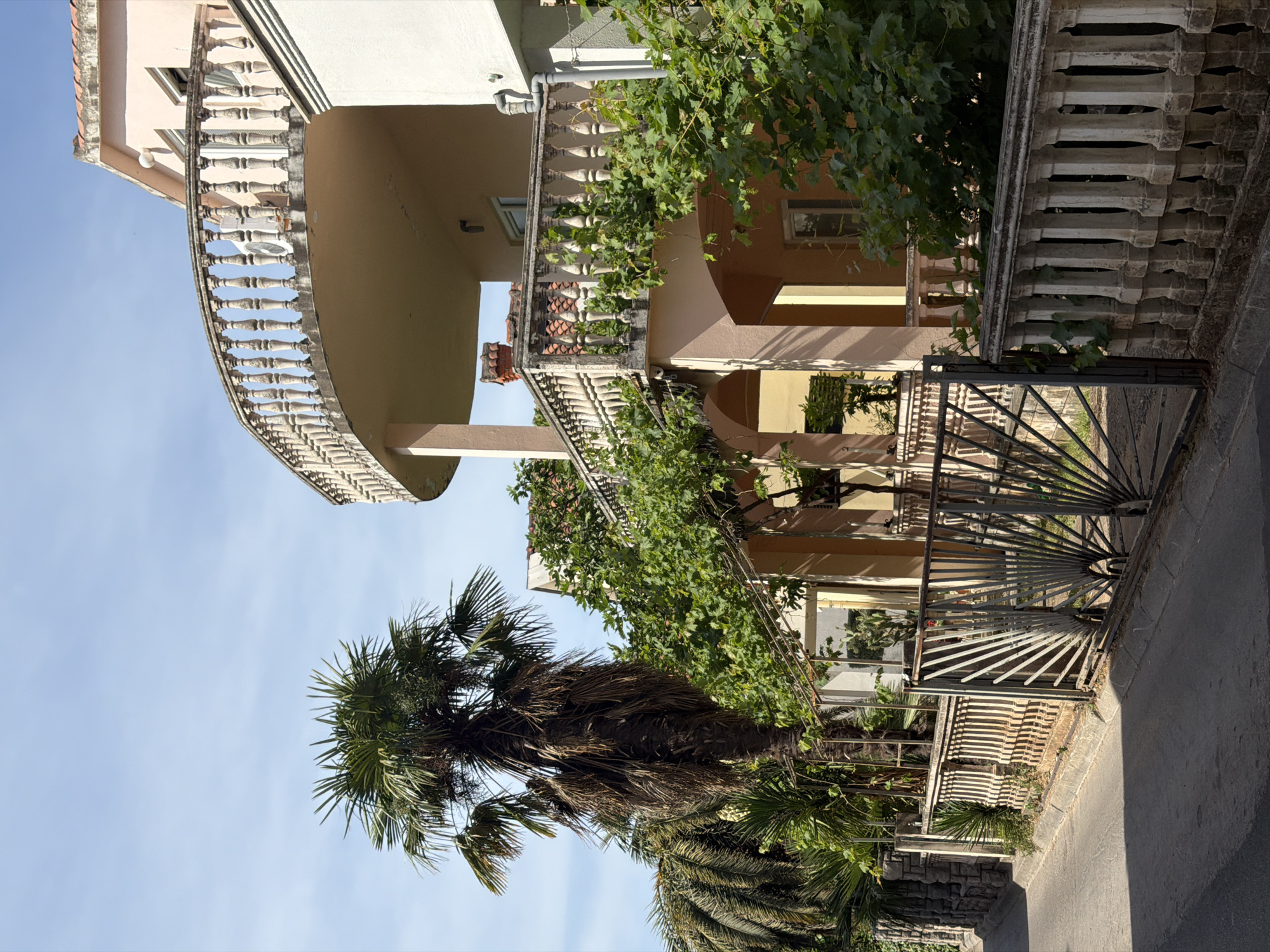 The image shows a view of a building in Budva, Montenegro. The building has multiple levels, each with a balcony featuring ornate balustrades. Lush green vines and other greenery adorn the balconies and facade. A palm tree stands in the foreground, partially obscuring the building. The sky is light blue with no clouds. A metal gate is visible near the bottom of the image, leading onto a paved road. The architectural style suggests a Mediterranean influence.