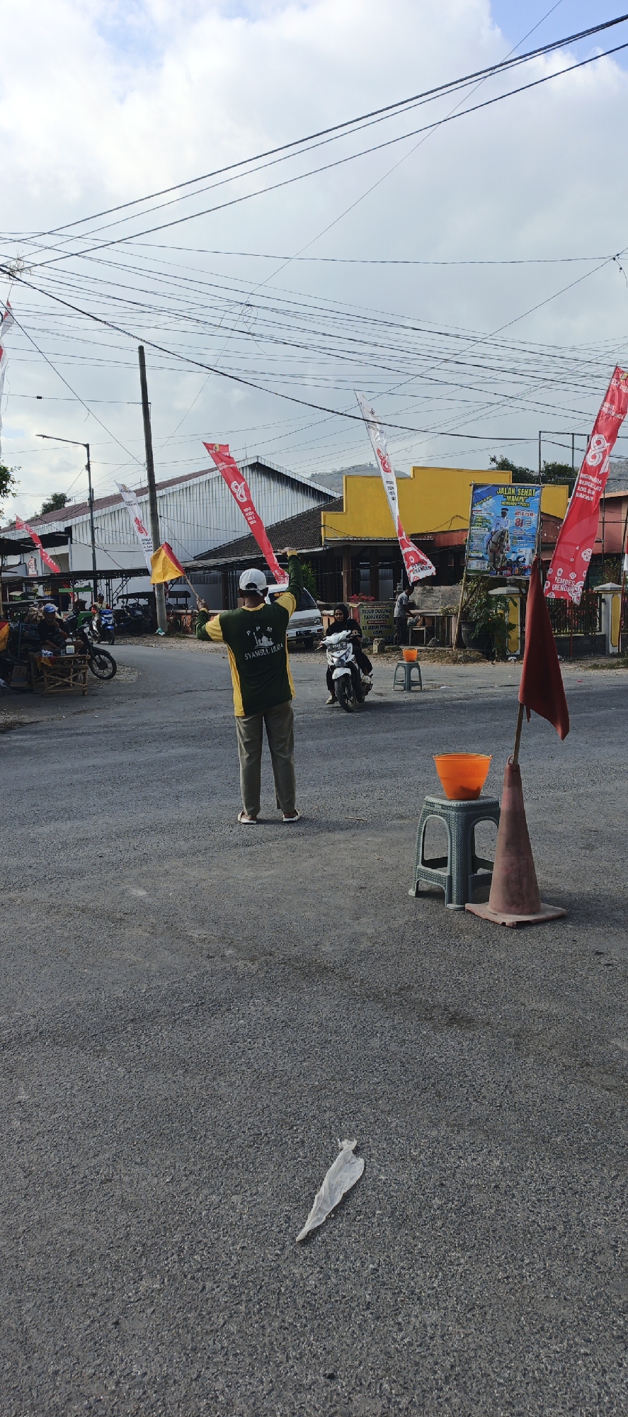 In the city of Grenden, Indonesia, a person stands in the middle of a road, holding two flags. One flag is red and white, and the other is yellow. The person is wearing a green and yellow shirt, khaki pants, and a white cap. They are facing away from the camera, directing traffic with the flags. To the right of the person, a red traffic cone and a small green stool with an orange bucket on top are visible. A motorcycle passes by in the background. Buildings line the street, and overhead power lines crisscross the sky. The sky is partly cloudy. A white plastic bag lies on the asphalt in the foreground.