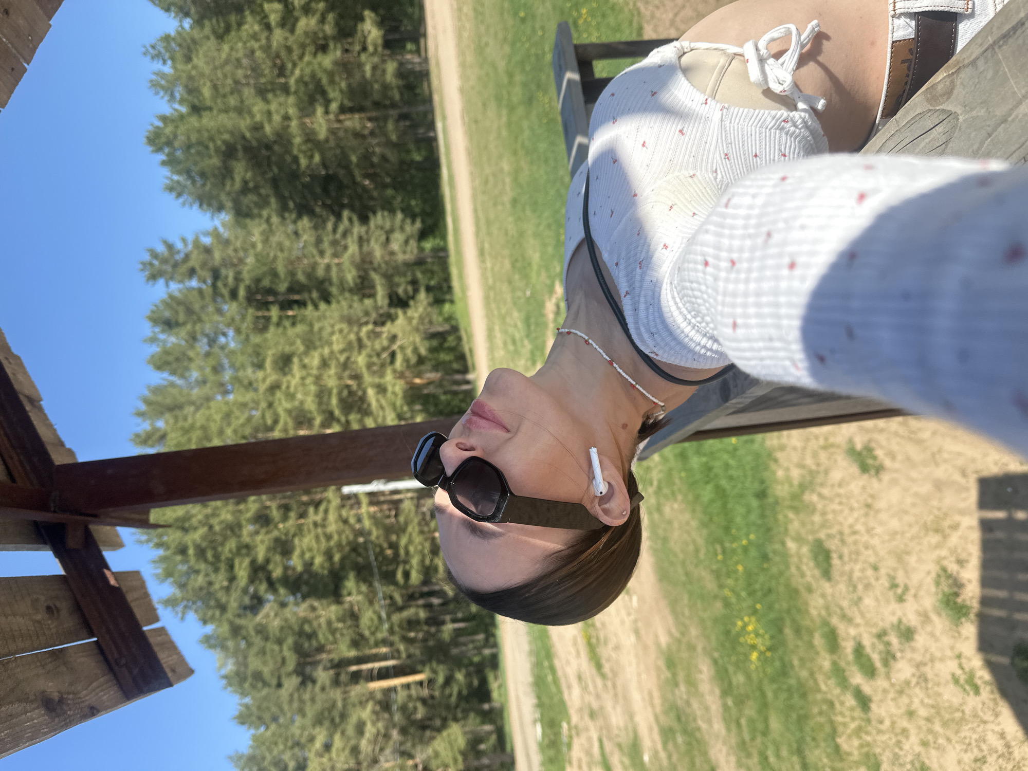 A young woman wearing sunglasses and an earpiece leans back on a wooden structure, potentially a gazebo or observation tower. She is wearing a white top with a red pattern. Trees and a grassy area are visible in the background, suggesting a park or natural setting. The location is likely Votkinsk, Russia, based on the prompt. The sky is clear and blue.