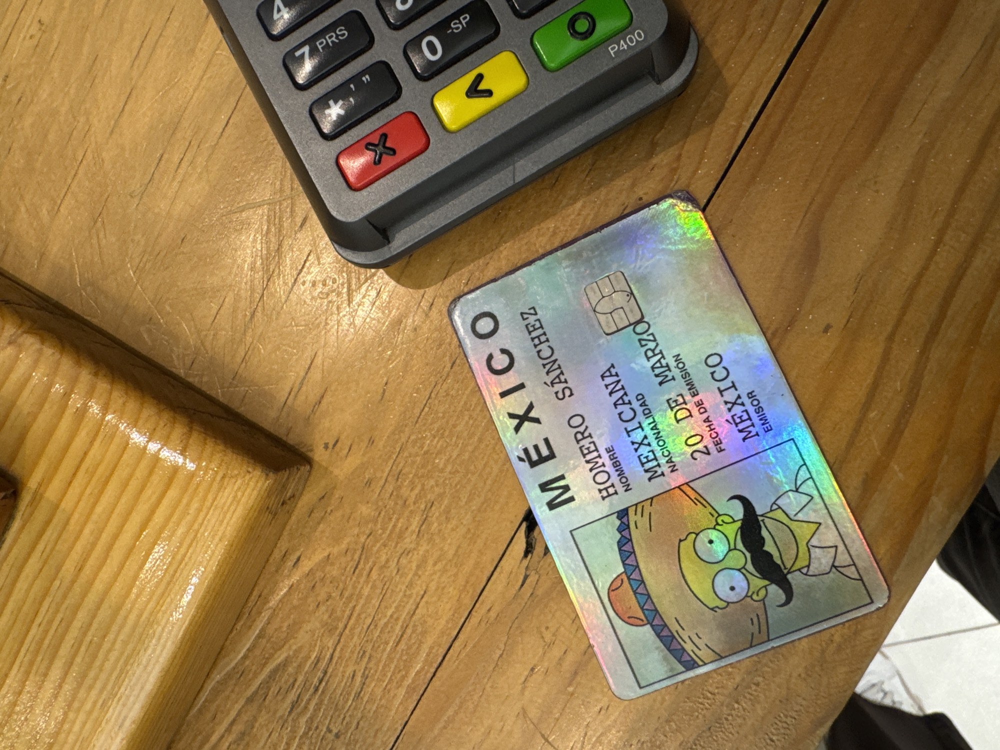A Mexican ID card featuring a caricature of Homer Simpson with a mustache is on a wooden surface next to a credit card terminal. The ID card reads "MEXICO" vertically in bold letters. The name "HOMERO SANCHEZ" appears beneath. The nationality is listed as "MEXICANA," and the date of issue is "20 DE MARZO." The issuer is "MEXICO." The credit card terminal has visible buttons, including numbers, a yellow ">" button, a green circle button, and a red "X" button. The wooden surface shows grain patterns.