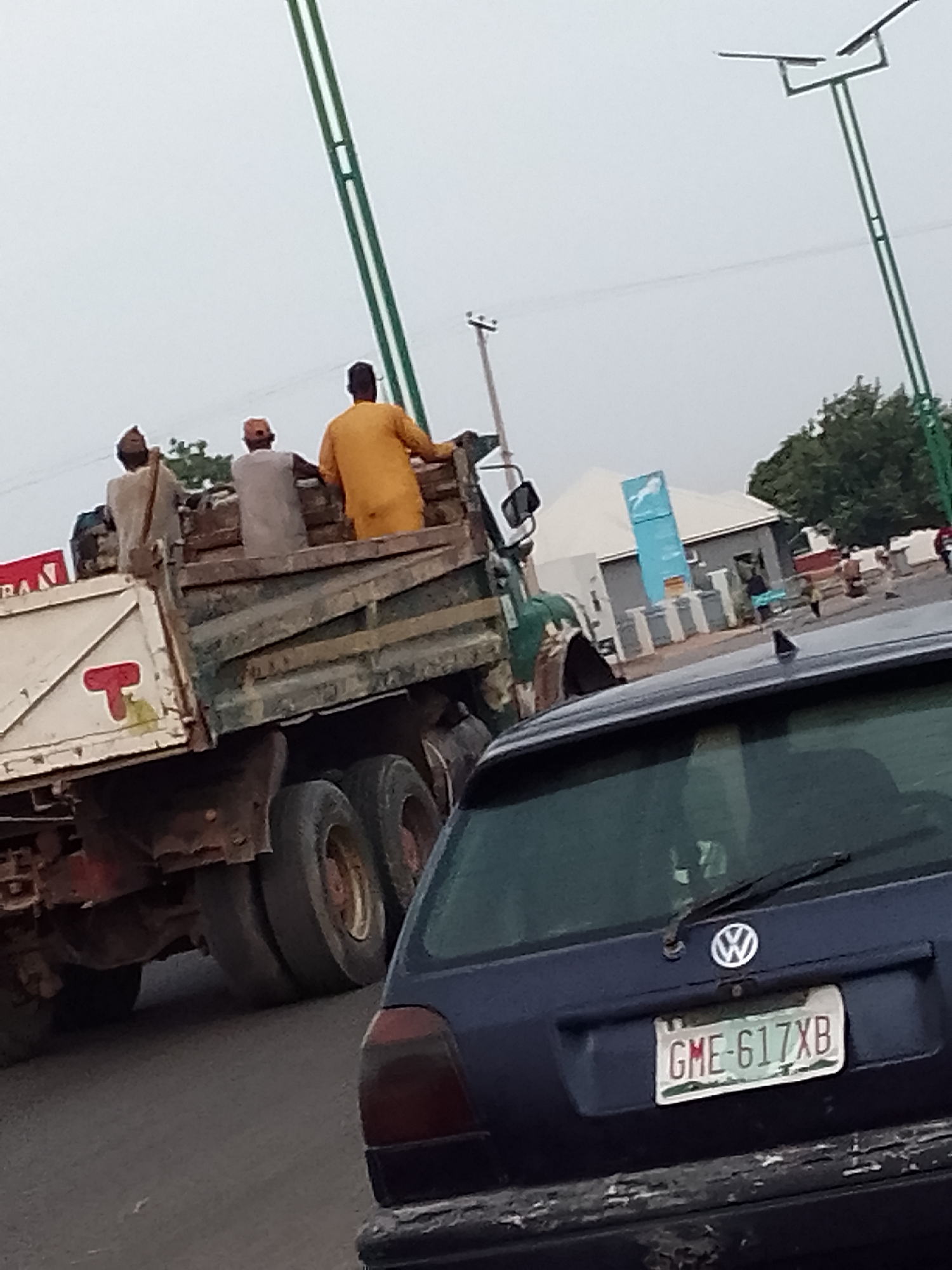 In Pantame, Nigeria, on an overcast day, a dark blue Volkswagen hatchback is parked in traffic, its rear bumper weathered. A license plate with the characters "GME-617XB" is affixed to the trunk. In front of it, a large dump truck, laden with dirt, is also in traffic. Three men, likely laborers, are seated in the open bed of the truck. One man in an orange garment faces away from the camera, while the other two are partially obscured. The truck's tires are worn, and its metal bed shows signs of age and use. Further in the background, a street lamp with solar panels stands tall, and a modern building with a blue sign is visible, suggesting a commercial or administrative area within the city. The road surface appears to be asphalt.