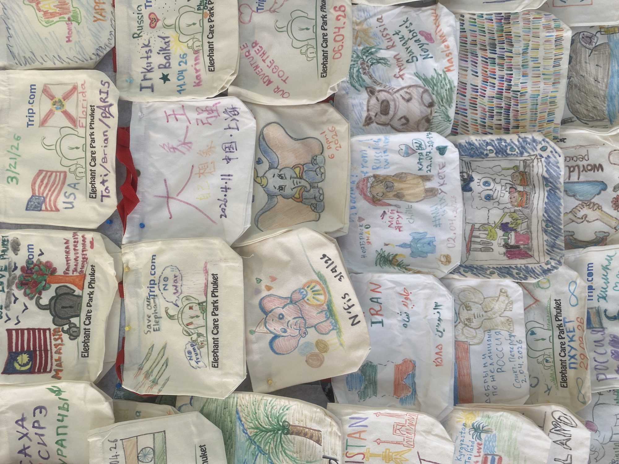 The image displays a vibrant collection of small, light-colored fabric bags, likely canvas or cotton, arranged and pinned together on a flat surface, forming a dense, colorful mosaic. Each bag serves as a personalized canvas, adorned with unique drawings and handwritten messages.
The subjects depicted vary, with elephants being a recurring motif, often drawn in cute, stylized, or celebratory ways. Other illustrations include national flags (USA, Malaysia, India, Russia, Iran), cartoon characters (Dumbo, Spongebob Squarepants), various animals (hippos, cats), and natural elements like palm trees and flowers. The artistic styles range from simple, childlike doodles to more intricate designs.
The bags are part of a public display at the Elephant Care Park Phuket in Choeng Thale, Thailand. The activity involves visitors or participants creating and contributing these decorated bags. The content of the bags suggests a focus on compassion, global unity, and advocacy. Visible messages include calls for "World Peace," "Save our Elephants," and "No War," with some expressing political views like "No Trump." Many bags feature names, personal greetings, and locations from around the world such as "USA," "Florida," "Paris," "Irkutsk Russia," "China," "Shanghai," "Malaysia," "Iran," "Kyrgyzstan," and various Russian cities. Numerous dates are prominently displayed, with many indicating "2026" (e.g., 3/21/26, 06.04.26, 6 April 2026, 2026.4.11), suggesting a forward-looking project or an accumulation of submissions over time, alongside older dates like 2017 and 2016. The visible text also includes website mentions like "Trip.com" and clear identification of the "Elephant Care Park Phuket." The overall impression is one of international engagement, shared values for peace, and support for elephant welfare.