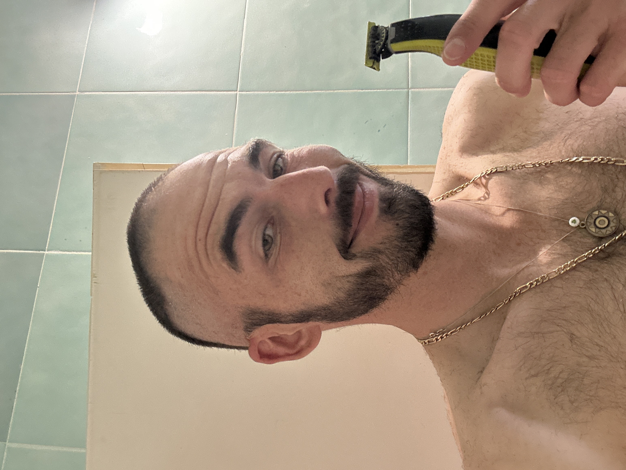 A man is holding a black and yellow electric razor to his chest. He has a beard and mustache and is wearing a gold chain necklace. The background appears to be a bathroom with light green tiled walls. It is not possible to verify the location in the city of Budva, Montenegro from the image.