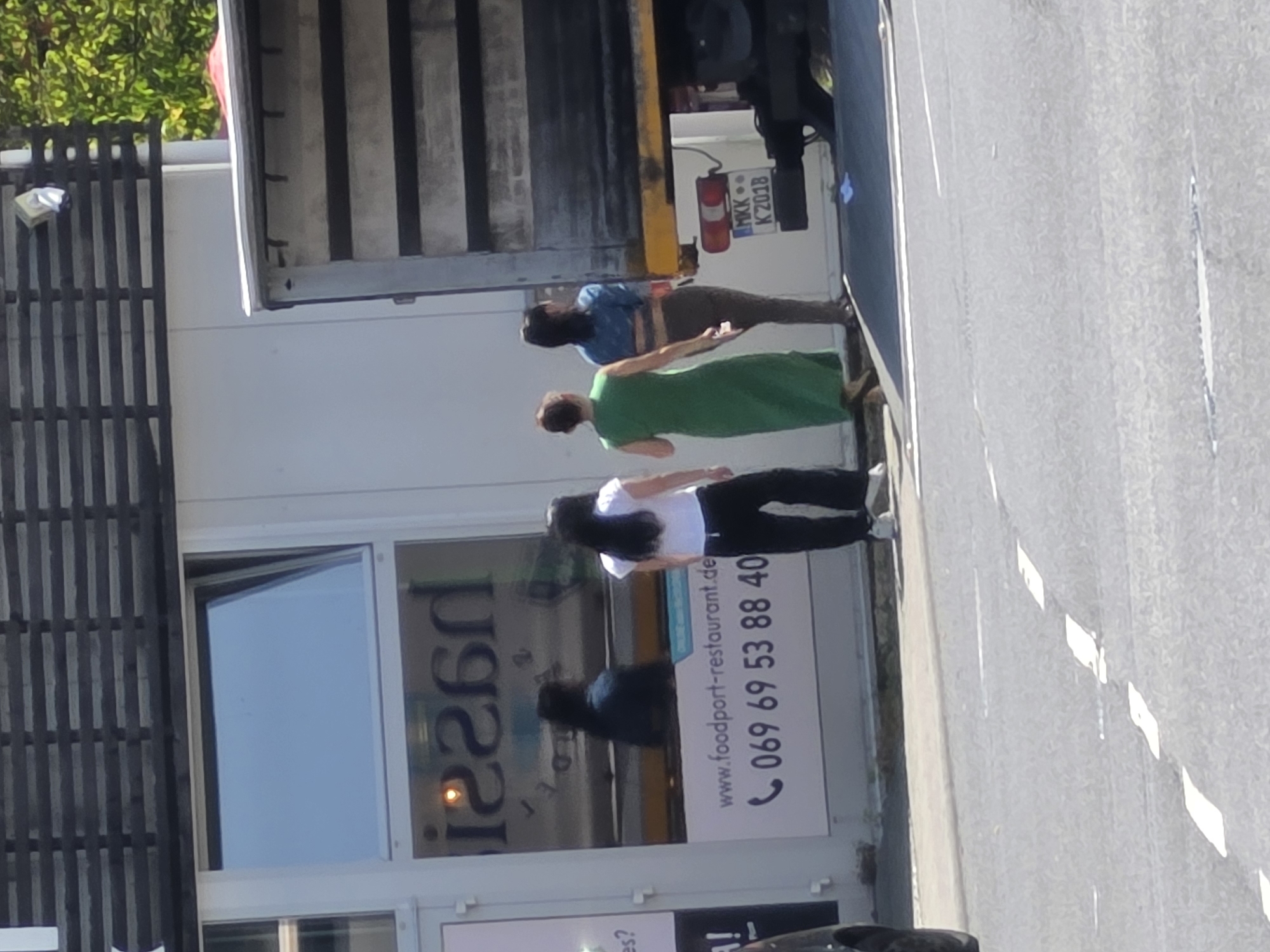 In Zeppelinheim, Germany, on a sunny day, three women are standing near a parked truck. The woman on the left wears a white t-shirt and black pants. The middle woman is in a long green dress. The woman on the right wears a blue jacket and dark pants. The truck, parked next to a building with a large window displaying a restaurant logo and website, has a license plate that reads "MKK K 2018". The building appears to be a commercial establishment, possibly a restaurant given the signage. The road in front of the building has white lane markings.