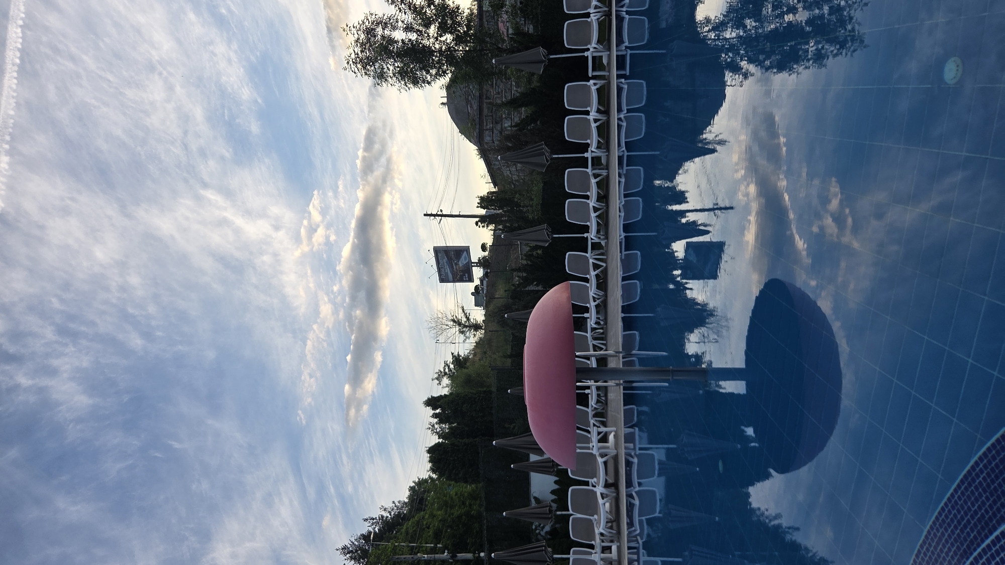 In the city of Unknown, Unknown, an outdoor swimming pool area is captured at dusk. The sky is a canvas of soft blues and whites, with wispy clouds stretching across the horizon, illuminated by the fading sunlight. A prominent pink mushroom-shaped structure stands in the foreground, partially submerged in the clear blue water. Rows of empty white lounge chairs and closed umbrellas are neatly arranged on the poolside, suggesting a quiet, end-of-day scene. Lush green trees and vegetation line the background, with a billboard visible on a distant hill. The still water of the pool reflects the sky, the surrounding trees, and the poolside amenities, creating a serene and symmetrical composition. The overall atmosphere is peaceful and tranquil.