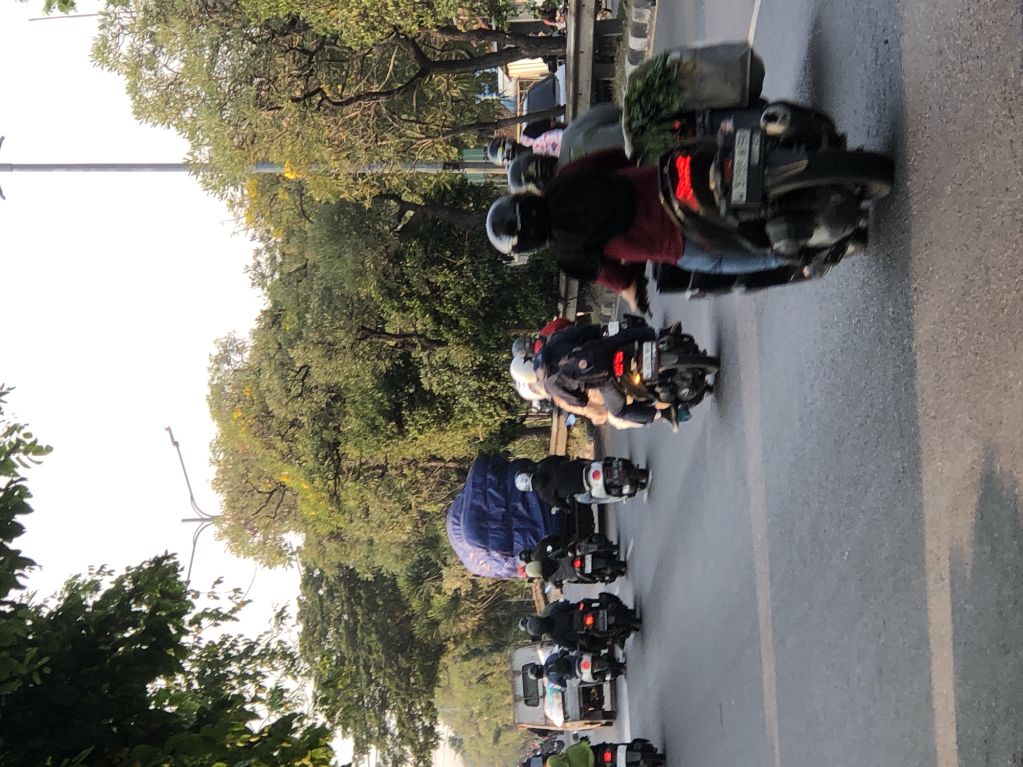 The image captures a group of motorcyclists riding on a paved road, likely in Kludan, Indonesia. The scene appears to be during the daytime, with the sky bright and some shadows visible on the road, suggesting it could be late afternoon. The weather seems to be clear, with lush green trees lining the roadside.
The primary subjects are the motorcyclists, most of whom are wearing helmets for safety. They are riding in a line, indicating they are part of a convoy or simply traveling together. The motorcycles are varied, with some appearing to be scooters or smaller displacement bikes. One motorcycle in the foreground has a cargo carrier on the back, filled with what looks like greenery. Another motorcyclist is wearing a maroon jacket and dark pants, while others are in darker attire.
The setting is a road surrounded by abundant foliage, with tall trees and bushes. A utility pole or street light is visible on the left side of the image. The road has lane markings, and there is a slight wetness on the surface, which might indicate recent rain or morning dew.
The activity is clearly motorcycle riding. The composition suggests movement, with the subjects captured in mid-journey. There are no overt signs of specific emotions, but the general atmosphere appears to be that of everyday travel.
There is some visible text on the license plate of one of the motorcycles, which appears to be a sequence of letters and numbers, but it is too blurred to read clearly. The overall impression is a common sight of motor vehicle traffic in a tropical or subtropical environment.