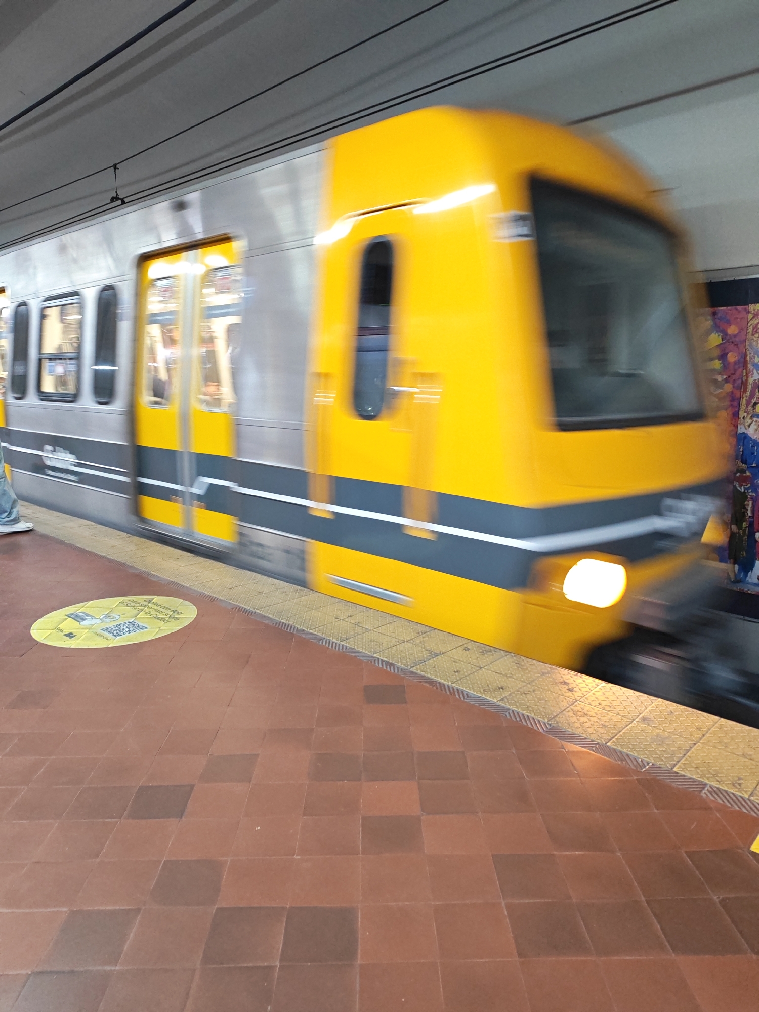 A subway train is in motion at a subway station in Buenos Aires, Argentina. The train is silver and yellow, with a dark gray stripe along the bottom. The front of the train is yellow and is moving rapidly past the viewer. The platform is made of brown and reddish-brown square tiles. The edge of the platform is lined with a yellow textured safety strip. There is a circular sign on the platform with yellow and white writing. In the background, a colorful mural is partially visible behind the moving train.
