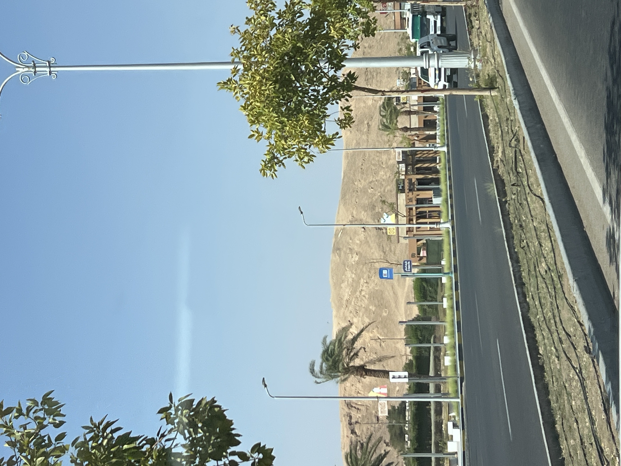 The image depicts a roadside scene in Sharm El Sheikh, Egypt, under a clear blue sky. A paved road runs horizontally across the lower portion of the frame, with distinct white lane markings. A concrete curb separates the road from a strip of dry grass and some visible irrigation tubing.
Beyond the road, a pedestrian walkway is visible, bordered by a white railing and interspersed with lampposts and small trees. In the mid-ground, several buildings with awnings and outdoor seating areas are situated. A bus stop sign is mounted on a pole. Palm trees are scattered throughout the landscape.
In the background, a large, barren, sandy hill dominates the horizon. The colors are muted, with the browns and tans of the desert landscape contrasting with the green of the trees and the blue of the sky. A few vehicles, including a white SUV and a white bus, are traveling on the road. No people are clearly visible in the scene. The overall impression is one of a sunny day in a desert city, with infrastructure designed to manage the climate.