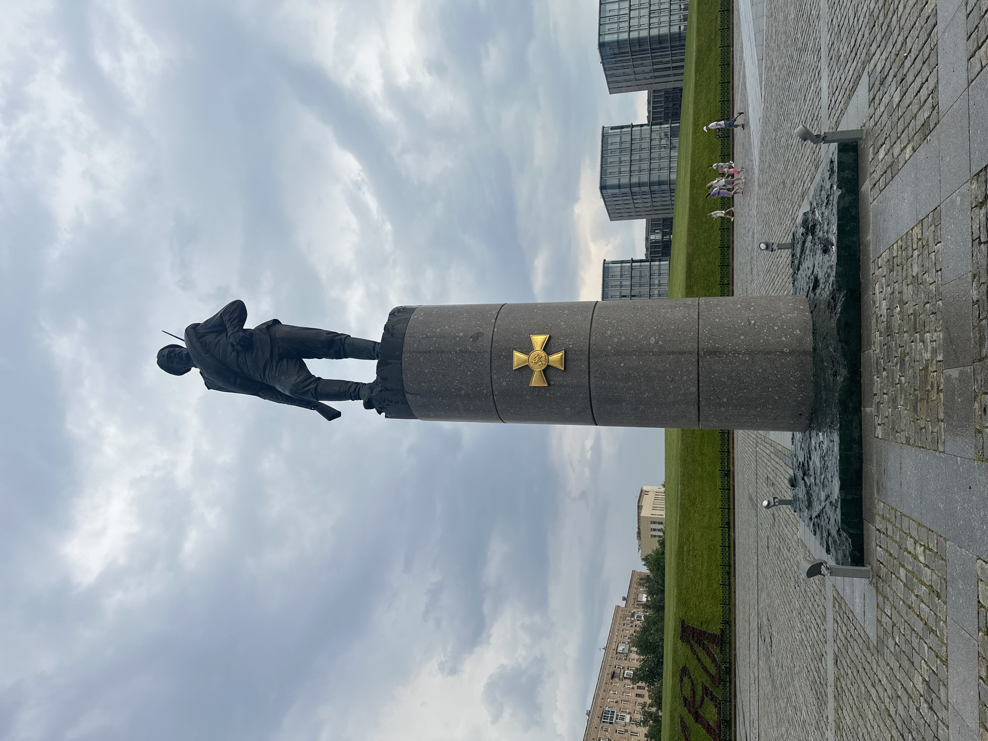 The image depicts a solemn statue of a soldier standing on a tall, cylindrical pedestal in a public square in Moscow, Russia. The soldier, rendered in dark bronze, holds a rifle and wears military attire. Centered on the pedestal is a prominent gold cross. The square is paved with cobblestones, and several modern glass-fronted buildings are visible in the background, situated behind a grassy hill. A group of people, including adults and children, are walking in the mid-ground. The sky is overcast with heavy clouds. The monument is located in the Museum of the Great Patriotic War on Poklonnaya Hill, Moscow.