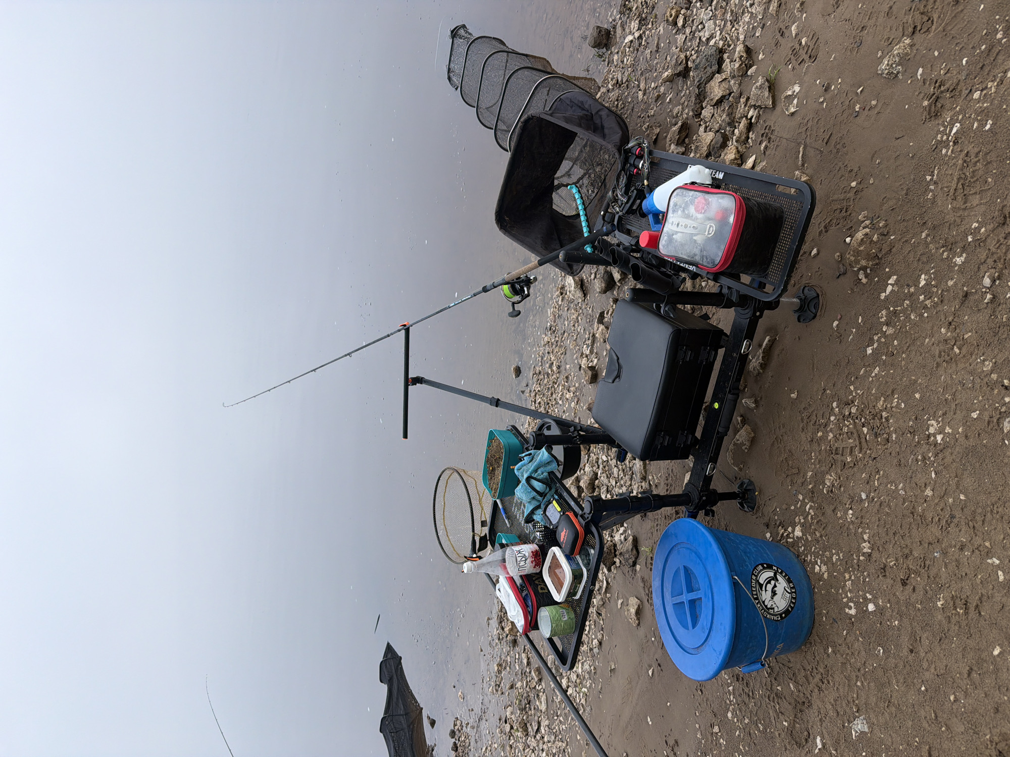 This media file captures a fishing setup on a muddy and rocky bank next to a calm body of water, likely a river or lake, under an overcast sky. No people are visible in the image.
The central subject is a black fishing station or seat, equipped with various fishing accessories. A fishing rod with a reel is positioned on a stand, its tip extending over the water. To the left of the station, a black keepnet is deployed into the water.
On the fishing station's side tray, a white bottle prominently labeled "ВОДКА КЕДРОВАЯ" (Vodka Kedrovaya) is visible, alongside several containers which likely hold bait or groundbait. One container has the partial text "DREAM FISH". A small fishing net and a blue towel are also on the tray. A red and black storage bag sits on the main platform of the station, which also has "TEAM" visible on its frame. Beside the station on the muddy ground is a blue bucket with a lid, featuring the text "BILLA FISHER TEAM" and "FEEDER CLUB" along with a fish logo.
The bank consists of dark mud interspersed with light-colored pebbles and rocks, showing some footprints. The water surface is still and reflects the pale, featureless sky, indicating an overcast day, possibly with misty conditions over the water. Further out in the water on the left, an indistinct dark shape might be additional fishing equipment. The scene depicts an ongoing fishing activity in an outdoor natural setting, potentially in Noviy, Russia, given the contextual information and visible text.