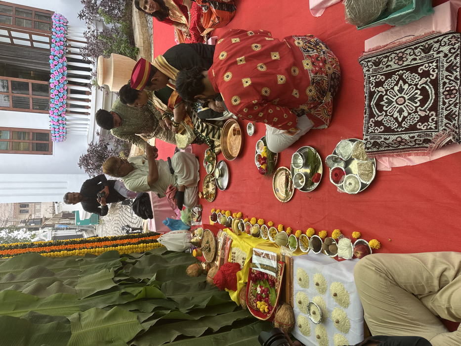 A group of people are gathered on a red carpet, seemingly participating in a traditional ceremony. Several metal plates holding items are arranged. A line of fruit and small bowls stretches across a low yellow table, while banana leaves line the bottom left. A white cloth with what appears to be rice and small silver bowls sits to the right. Several of the people are kneeling, while others stand or sit nearby.
