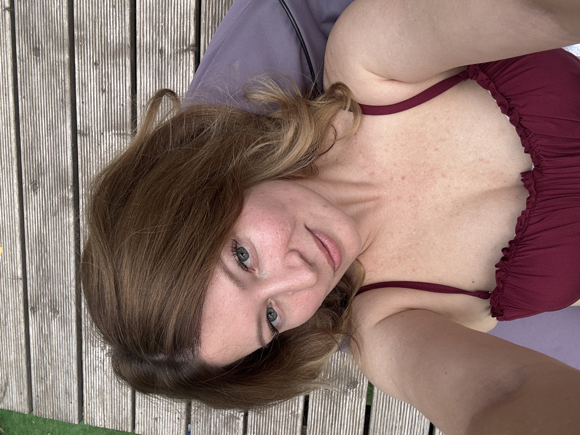 A woman is lying on a wooden deck next to a purple cushion. She is wearing a maroon bikini top. She has shoulder-length brown hair and green eyes. Her skin is fair with some redness on her cheeks and chest. The wooden deck is made of parallel planks. The background is out of focus but appears to be a grassy area. The image is a selfie, taken from a high angle, looking down at the woman.