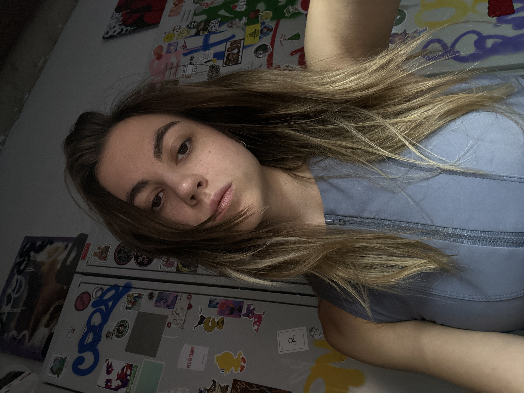 This is a selfie of a young woman with long, blonde hair. She is lying down, with her face angled towards the camera. She is wearing a light blue, zip-up top. Her hair is falling over her shoulders and chest. The background appears to be a wall with various stickers and posters, and what looks like a refrigerator covered in more stickers. Some of the stickers have text, but it is not legible enough to read clearly. The lighting suggests it is daytime, possibly indoors, as there are no strong shadows. The woman's expression is neutral, with her eyes open and looking directly at the camera. The overall impression is a casual, personal snapshot.
