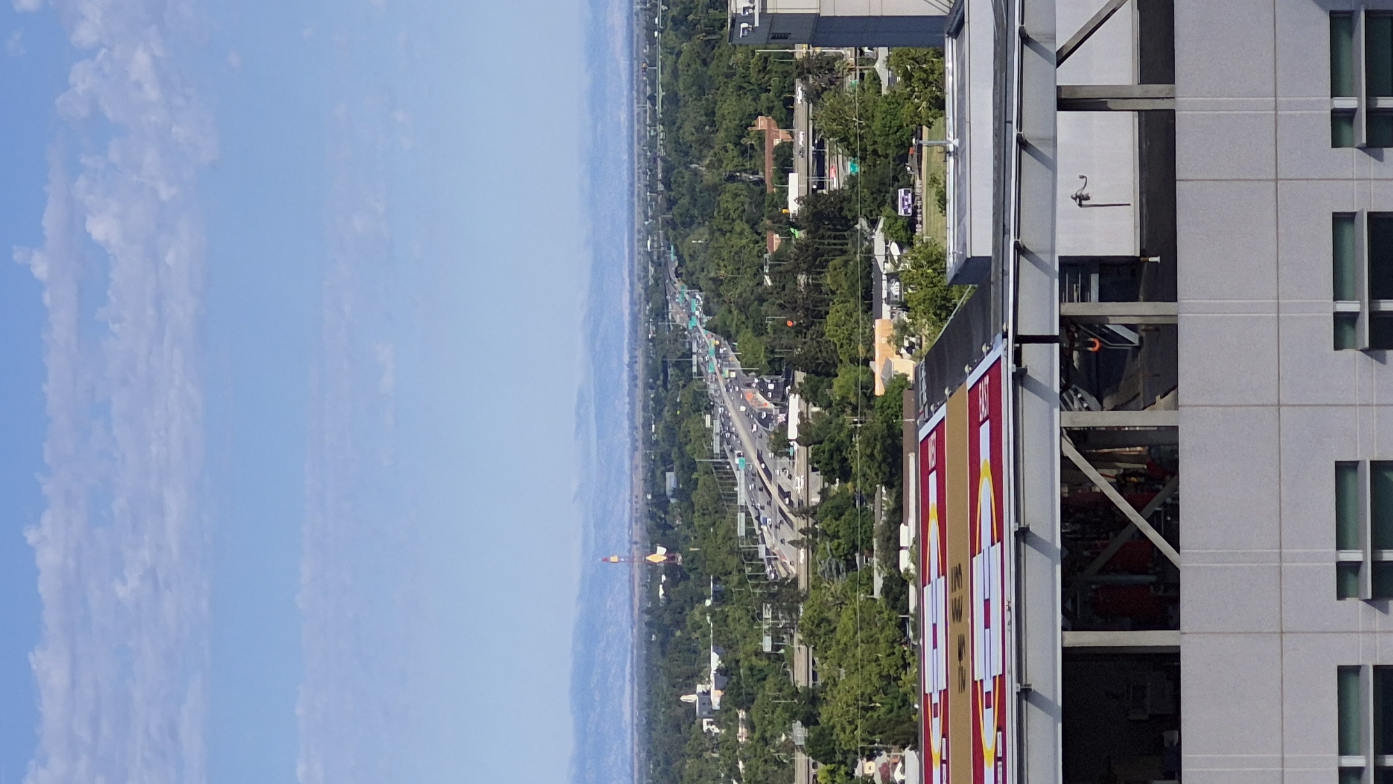 This is a vertical, eye-level, outdoor shot of a cityscape in Sacramento, California. In the foreground, a building with a white facade and multiple windows is visible. Above this building, a helipad with red, yellow, and white markings is seen. The middle ground of the image features a dense canopy of green trees and a highway with moderate traffic. In the distance, a tall tower with red and yellow bands stands out, and a mountain range is faintly visible on the horizon under a pale blue sky with scattered white clouds. The scene is captured on a clear day with ample daylight.
