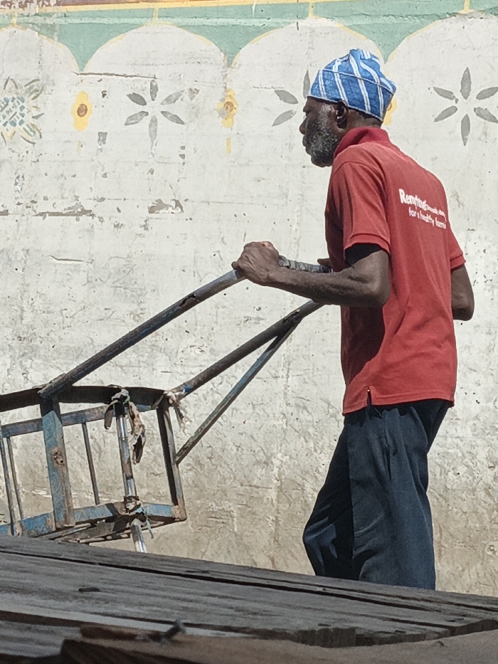 An adult male with dark skin and a graying beard, wearing a blue and white patterned skullcap, a red polo shirt with the text "Rentog... for a healthy home" on the back, and dark trousers, is depicted in Fagge C, Nigeria. He is actively pushing or pulling a worn, light-blue metal cart with visible rust and attached fabric scraps. His hands grip the cart's handlebars. The background consists of a weathered, light-colored wall adorned with a faded decorative pattern of arched segments and floral motifs in yellow and gray. Below the subject and cart, a dark, textured surface, likely wood or corrugated metal, occupies the foreground. The scene is brightly lit by natural sunlight.