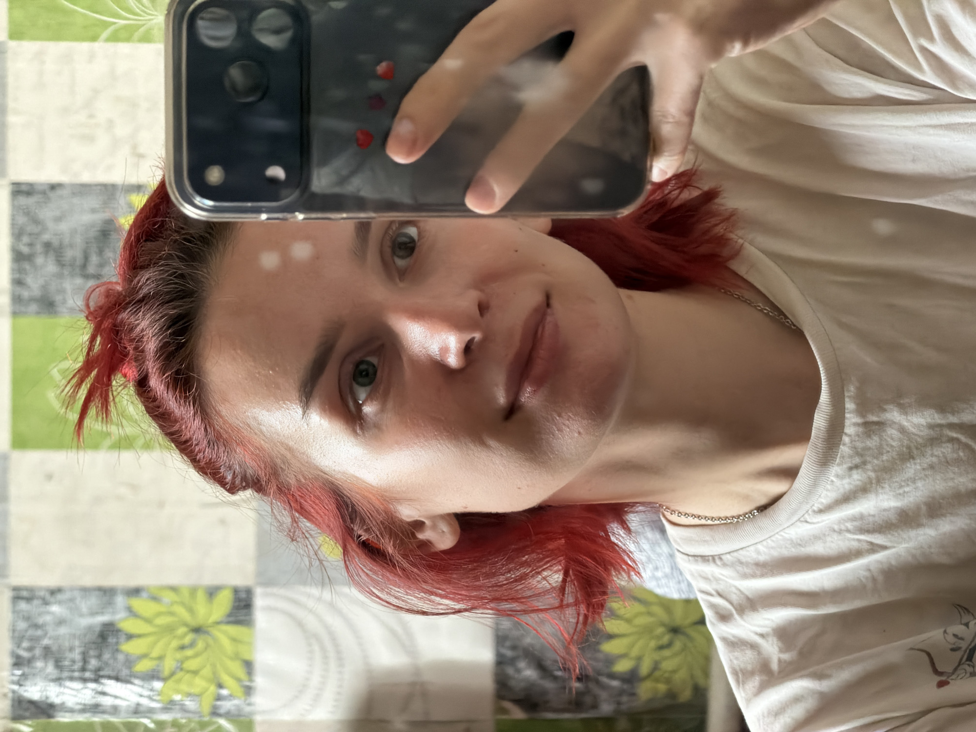 A woman with red hair is taking a selfie with a black smartphone in a clear case decorated with red hearts. She is wearing a white t-shirt and a silver chain necklace. The background is a table covered with a checkered tablecloth featuring green floral patterns. The location, Заболотово, Russia, is not discernible from the image.