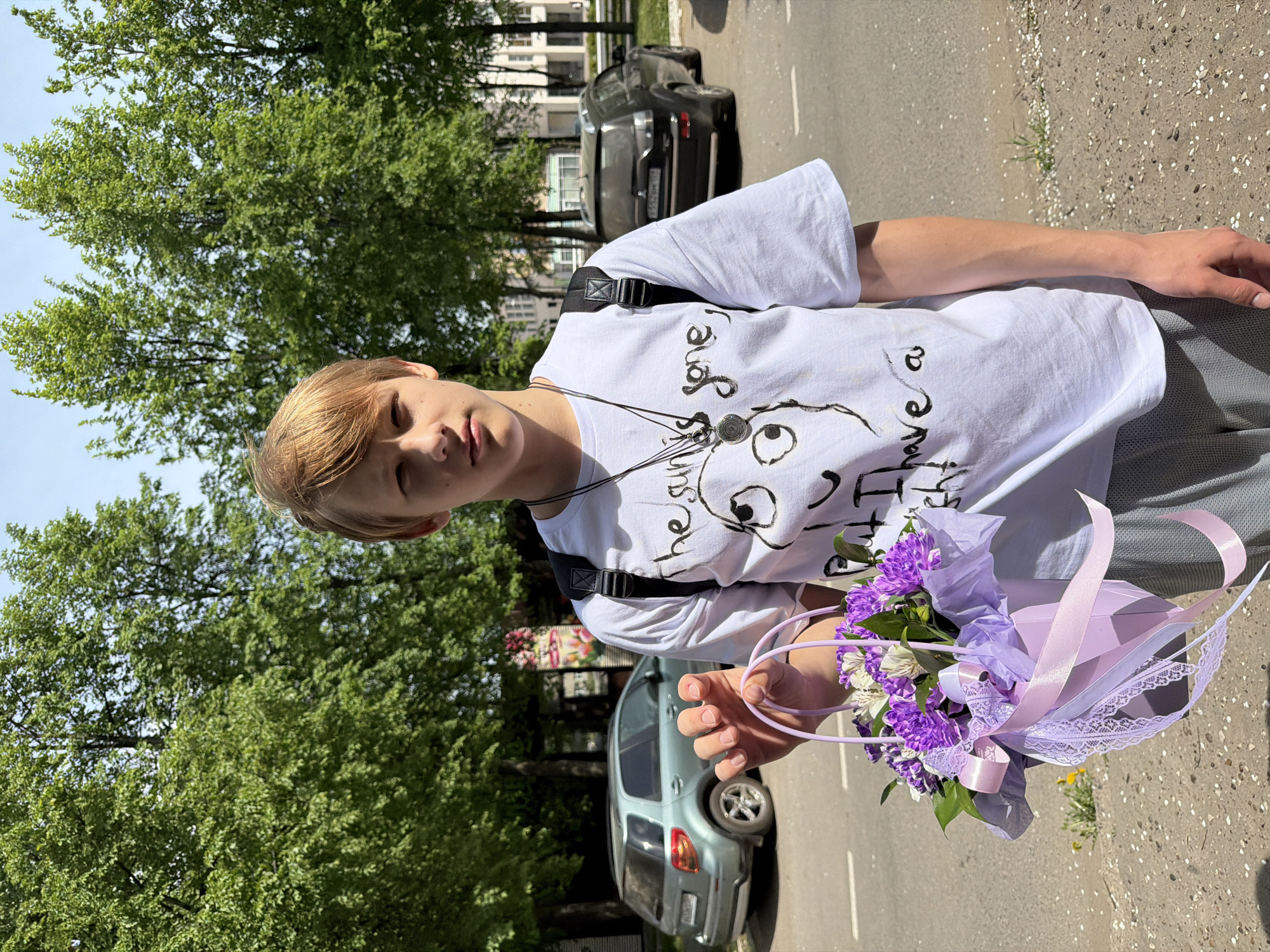 A young person stands on a sidewalk, holding a bouquet of purple and white flowers. The individual is wearing a white t-shirt with a black graphic and text, a necklace, and a black backpack. The background includes trees, cars parked on the side of the road, and buildings. The location is near a road in the city of Chaikovsky, Russia.