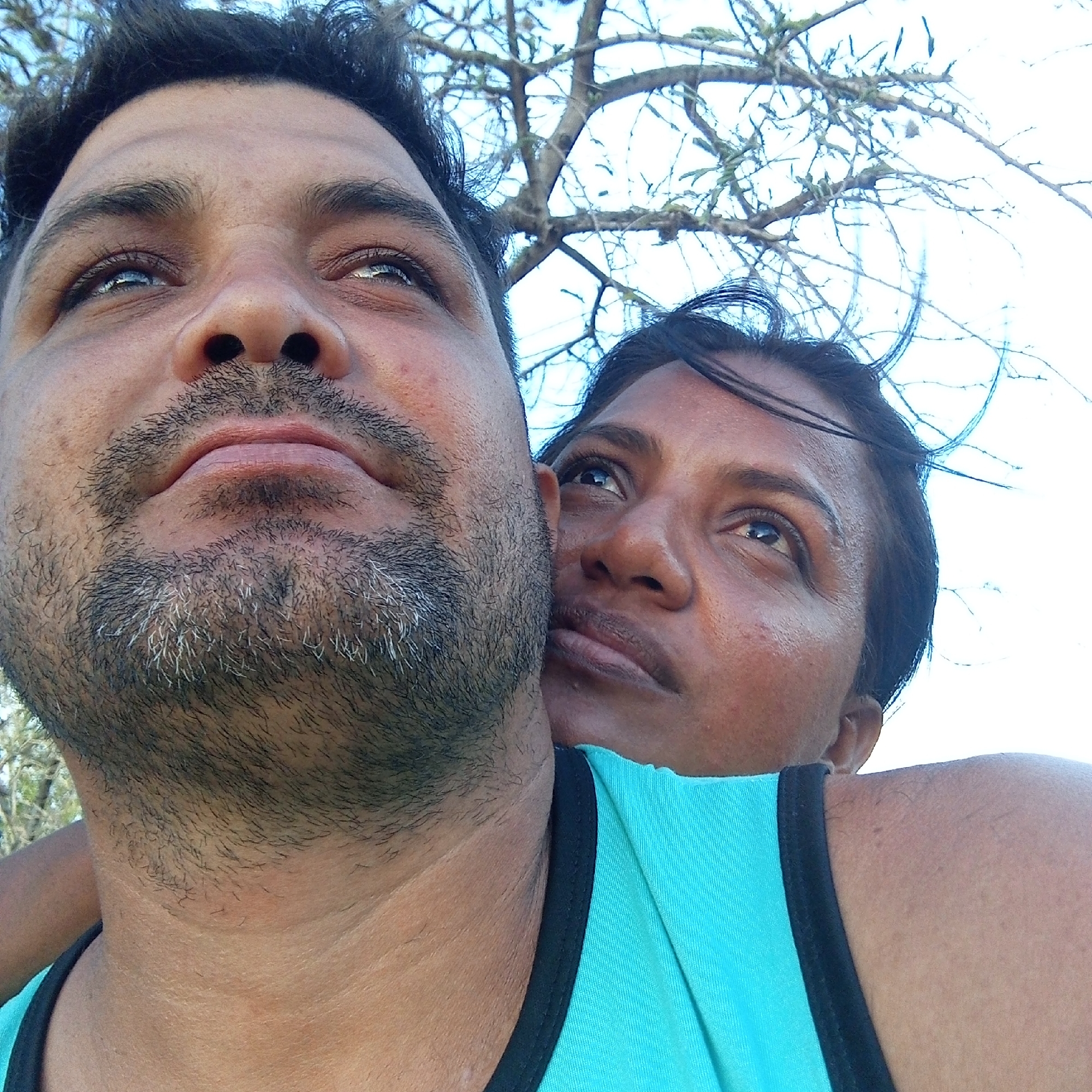 A close-up shot depicts a man and a woman looking upwards. The man, with dark hair and a beard, is positioned in the foreground. The woman, with dark hair pulled back, is slightly behind and to the side of the man, with her face visible behind his neck. Both individuals are fair-skinned and are wearing a light blue shirt with black trim. In the background, bare tree branches are visible against a bright sky. The location is unknown.