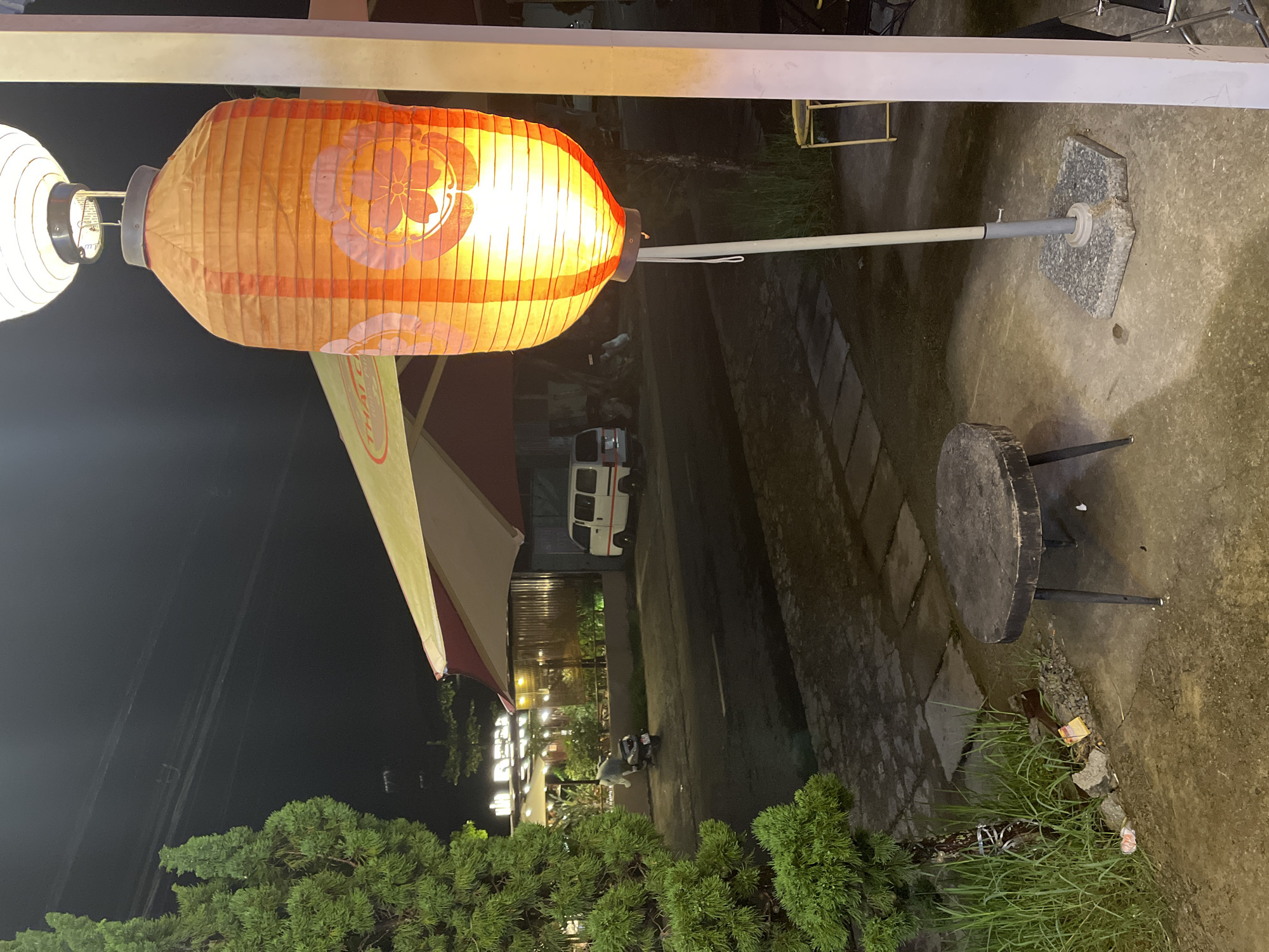 The image depicts an outdoor scene at night in Lâm Viên - Đà Lạt, Vietnam. In the foreground, a large, orange, cylindrical paper lantern hangs illuminated by an internal light source. The lantern has a floral pattern in a darker orange. It is suspended from a pole that is secured by a grey, stone base. To the left of the lantern is a small, round, dark wooden table with three metal legs. Adjacent to the table and extending to the left edge of the frame is a bushy green tree.
In the midground, a road is visible with a white van parked on the right side. Beyond the road, there is a building with a distinctive red and white awning. Lights are visible within the building, suggesting it is a commercial establishment, possibly a restaurant, as indicated by the visible signage which includes the word "THAI". A scooter is parked near the building on the left side of the road. The environment appears to be a street or a walkway adjacent to a street, with paving stones and some sparse vegetation. The lighting suggests it is nighttime, with artificial lights from the establishment and the lantern illuminating the scene.