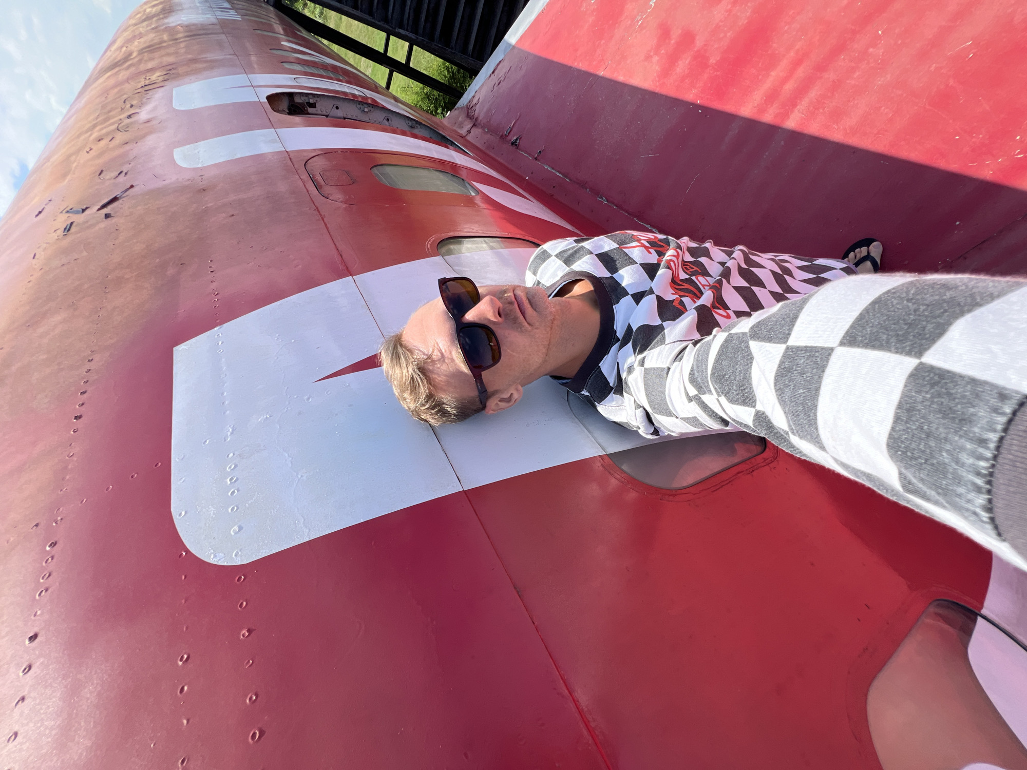 A man is lying on his back on the fuselage of a red and white airplane. He is wearing sunglasses and a black and white checkered long-sleeved shirt. The plane appears to be outdoors, with a blue sky and some green foliage visible in the background. The sun is shining, casting a shadow from the plane's structure onto the fuselage. The man is looking up at the sky, with a relaxed expression on his face. The airplane is large and cylindrical, with several windows visible along its length. The red and white paint scheme is bold and distinctive. The overall scene suggests a leisurely or adventurous moment, perhaps a tourist posing with a unique piece of aviation history. There is no visible text in the image.