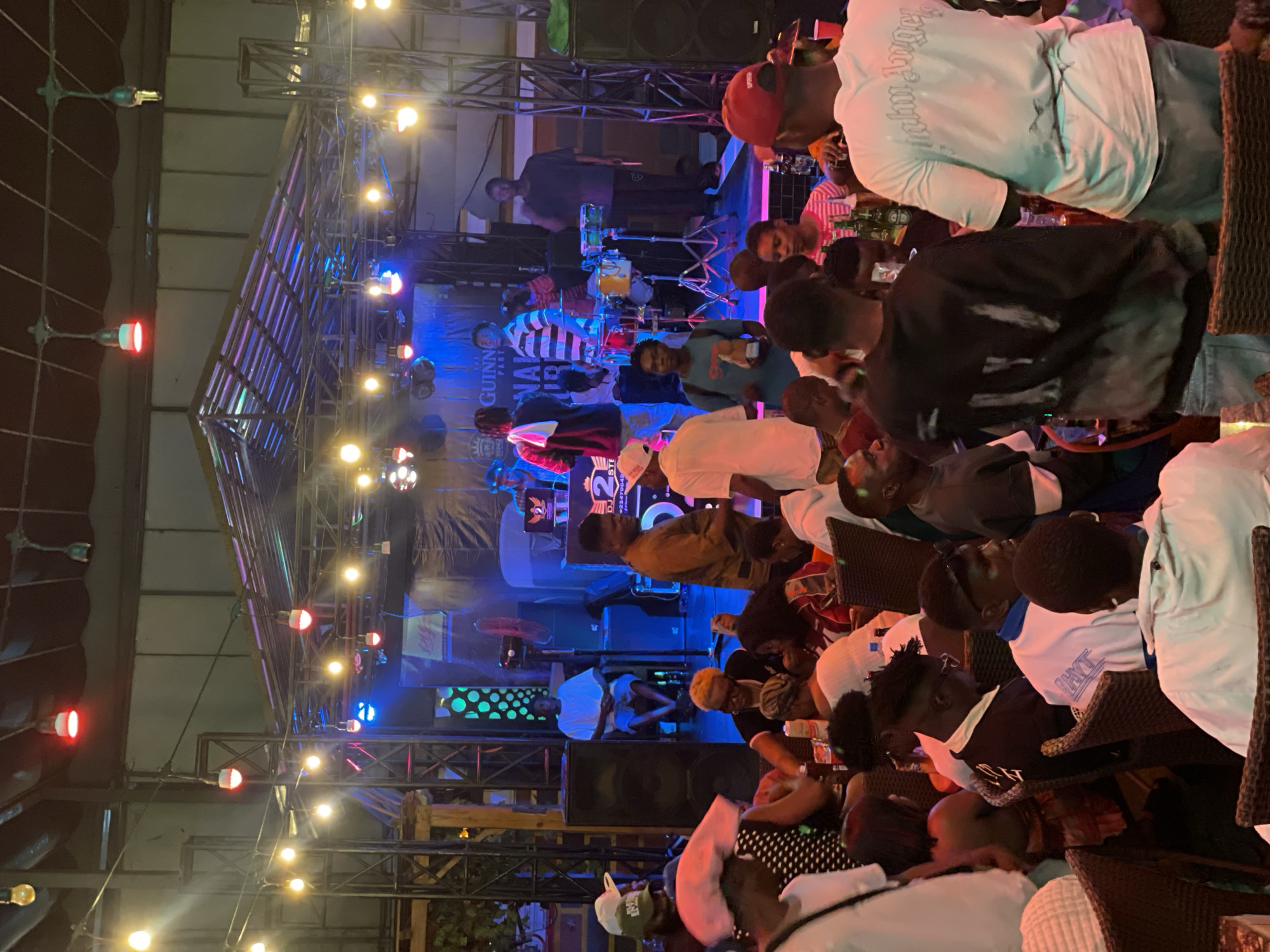The image depicts a live music performance at an outdoor venue in Obosi, Nigeria. A stage with metal scaffolding is illuminated with blue and yellow stage lights. A band is performing on stage, including a drummer. A crowd of people, primarily African, are seated at tables, watching the performance. Some individuals are wearing casual clothing. The venue is also decorated with hanging string lights.