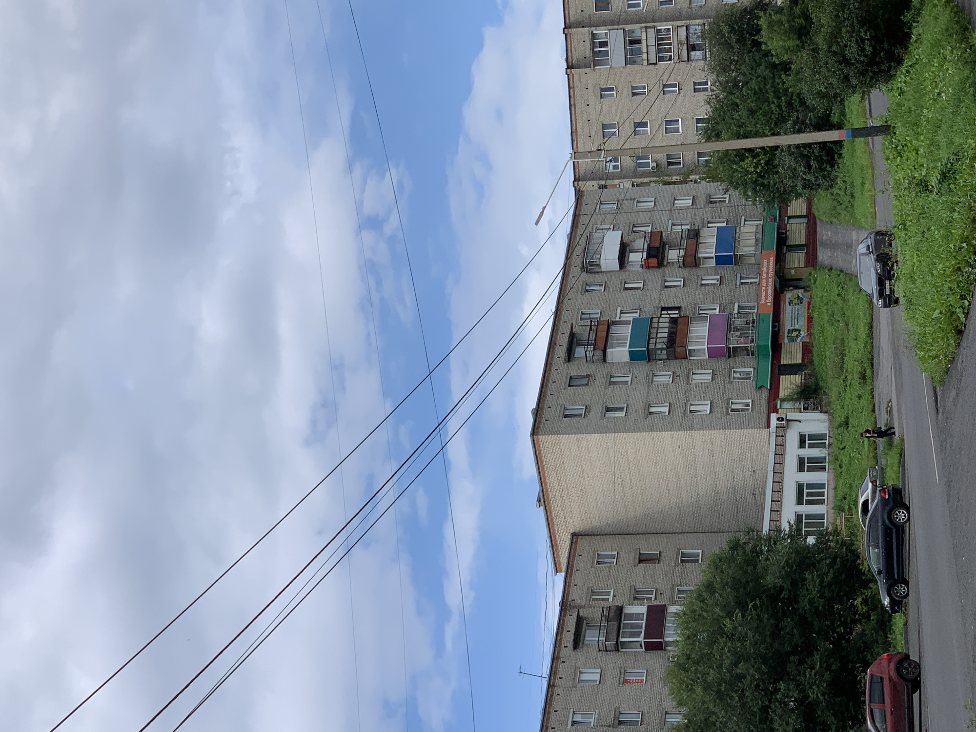 A street-level, eye-level, outdoor, natural-light photograph captures a cloudy day in Prokopyevsk, Russia. The scene is dominated by a long, multi-story apartment building, constructed primarily of light-colored brick. The building features numerous windows, balconies with varying railing designs, and what appears to be an advertisement for automotive parts on the ground floor. Power lines stretch across the upper portion of the frame, with the sky peeking through between them. The foreground shows a paved road and sidewalk, with several parked cars and a lone pedestrian walking. Lush green grass and trees surround the base of the building and line the roadside, indicating a verdant environment. The overall impression is of a typical residential street in a Russian city.