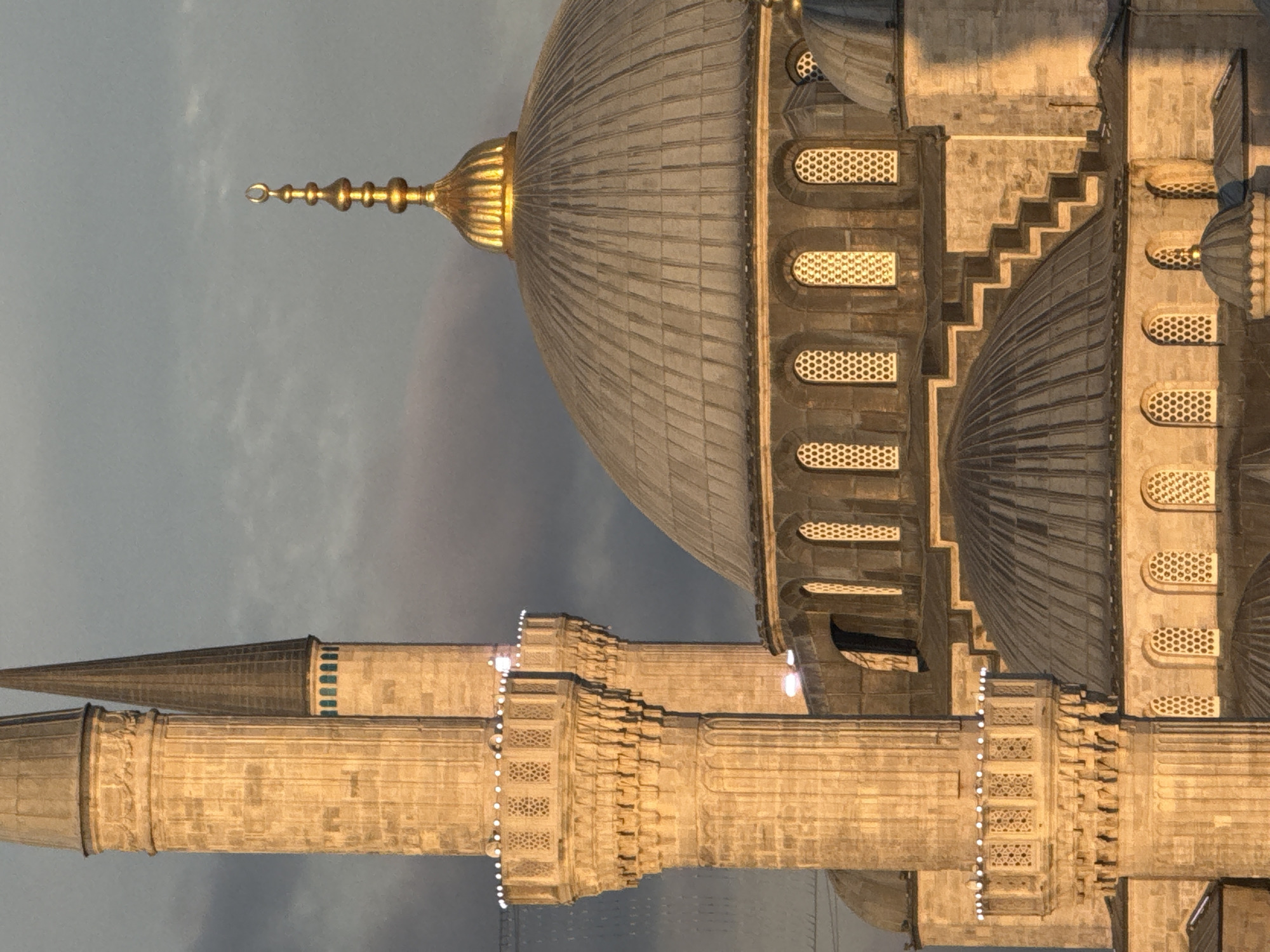 The image shows a close-up view of the Sultan Ahmed Mosque, also known as the Blue Mosque, located in Istanbul, Turkey. The mosque's distinctive minarets and domes are prominently featured, bathed in the warm, golden light of what appears to be either sunrise or sunset. The architectural details, including the stone carvings and decorative elements on the minarets, are clearly visible. The sky above is overcast with clouds, creating a dramatic backdrop. There are no people visible in the image. The focus is entirely on the magnificent structure of the mosque.