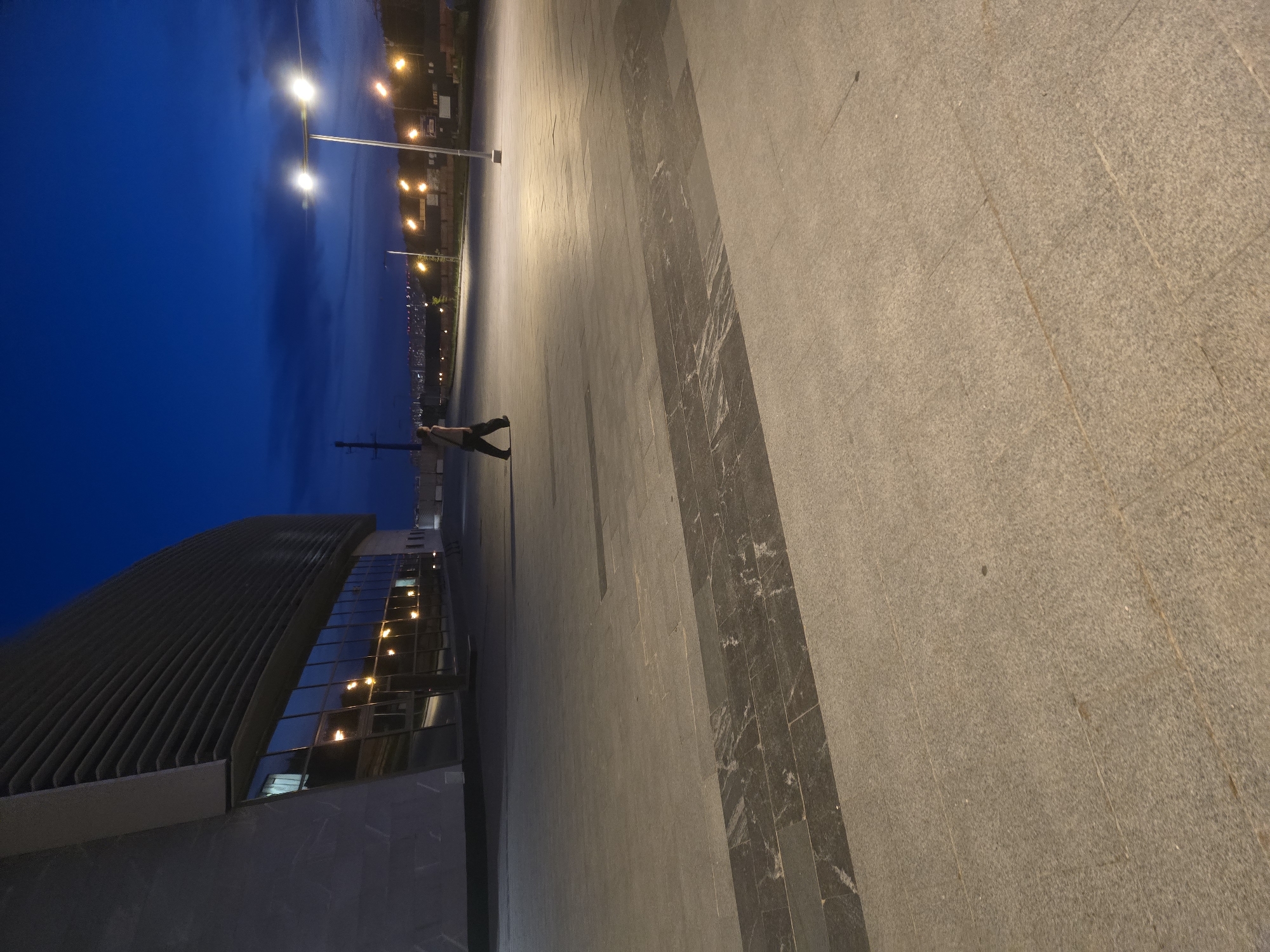 A full shot captures an outdoor scene at dusk, with a modern building on the left and a lone pedestrian walking across a paved plaza towards the right. The building, with a curved facade and rows of horizontal louvers, features large reflective windows that show interior lights. The plaza is paved with large, square tiles and a distinct rectangular section of darker, patterned tiles runs across the middle ground. Two streetlights illuminate the area, casting pools of light. In the background, more streetlights and distant city lights are visible against a darkening blue sky with faint clouds. The pedestrian, seen from the side, is wearing dark clothing and appears to be walking with a bag slung over one shoulder. The overall atmosphere is quiet and slightly somber due to the low light conditions.
